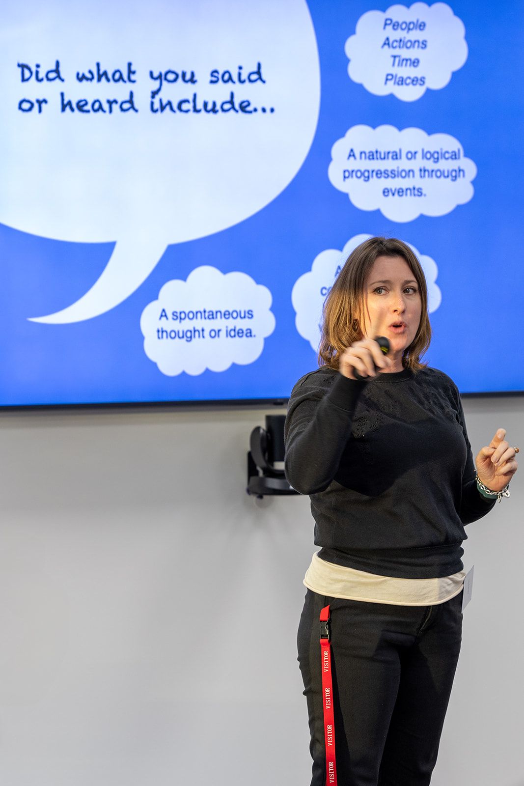 Woman presenter pointing at blue screen displaying speech bubbles with text about storytelling elements during a workshop.