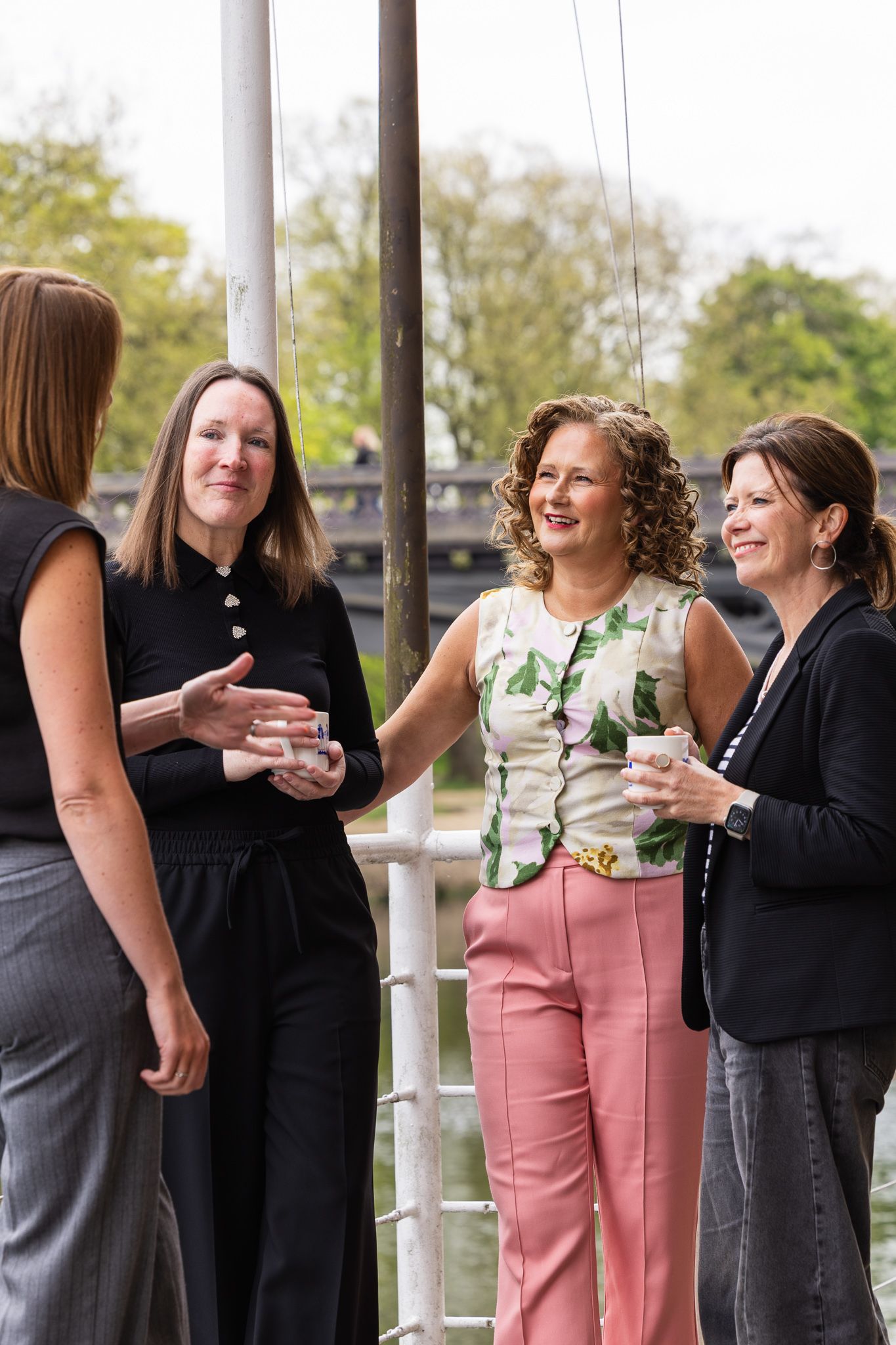 Four women socializing outdoors on a dock, smiling and holding drinks in a garden setting.