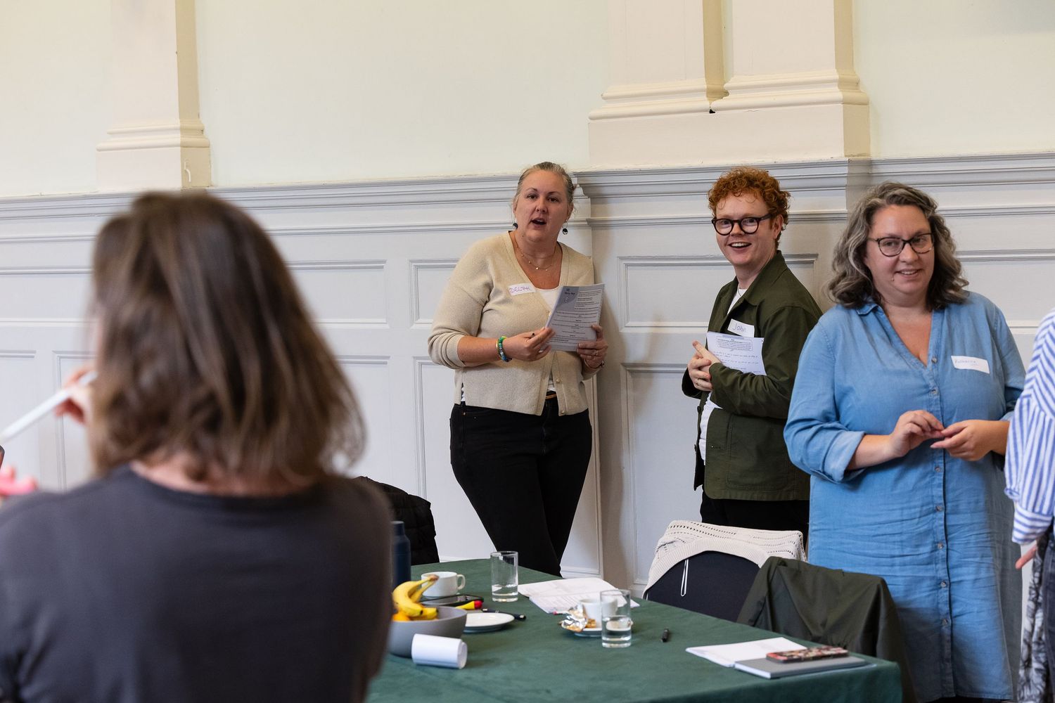 Three women stand near a green-draped table in a bright hall, one speaking while holding papers at a professional event.
