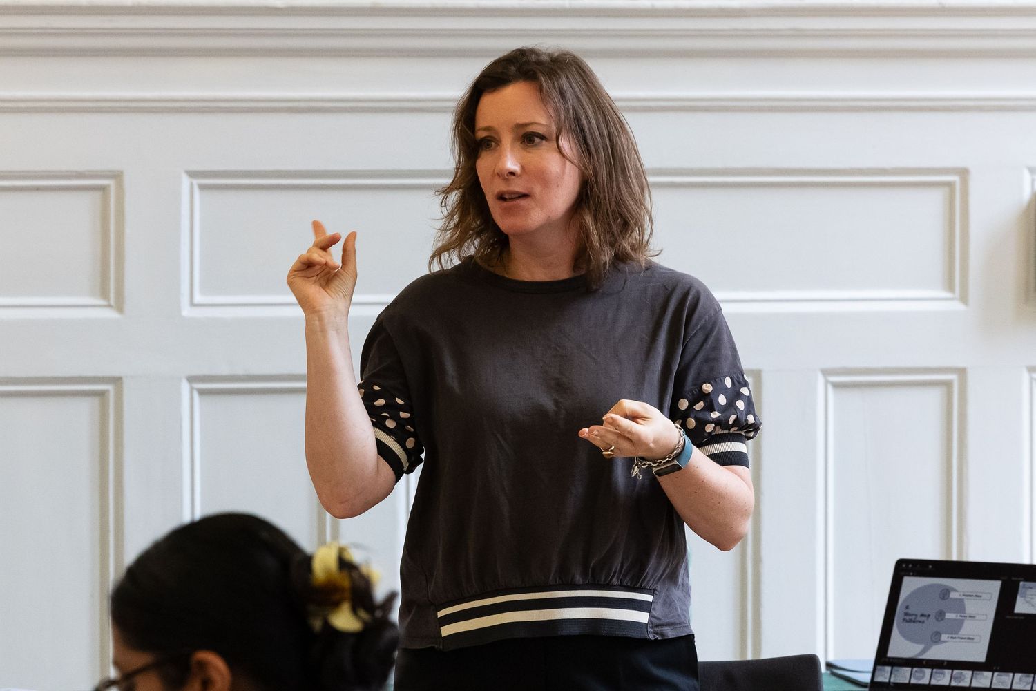 Woman in dark sweater gesturing while speaking at an indoor presentation or workshop setting.