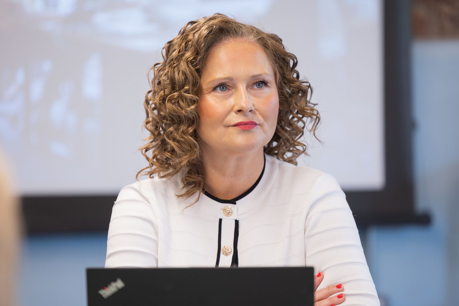 Professional woman with curly hair in white blazer seated at conference table with laptop.