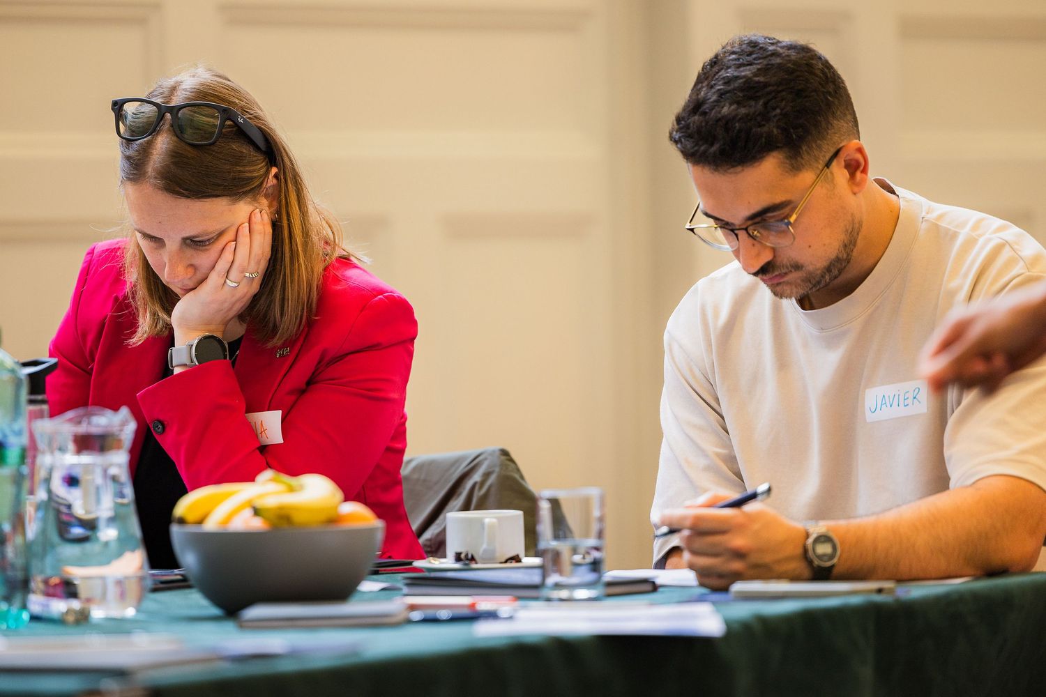 Two professionals focused on writing at a conference table with fruit bowl and glasses nearby.