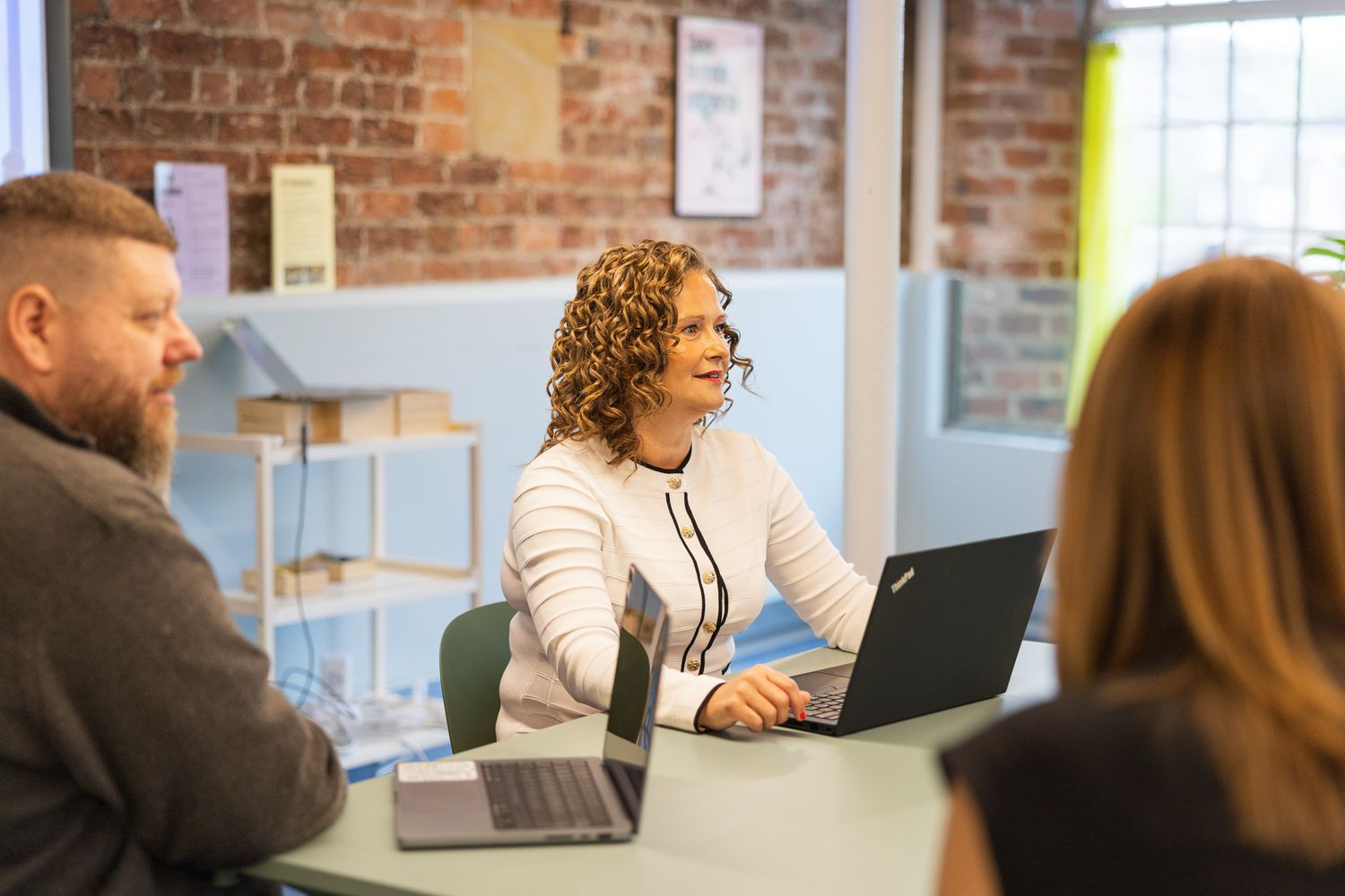 A woman with curly hair works on a laptop during a meeting in a modern brick-walled office.