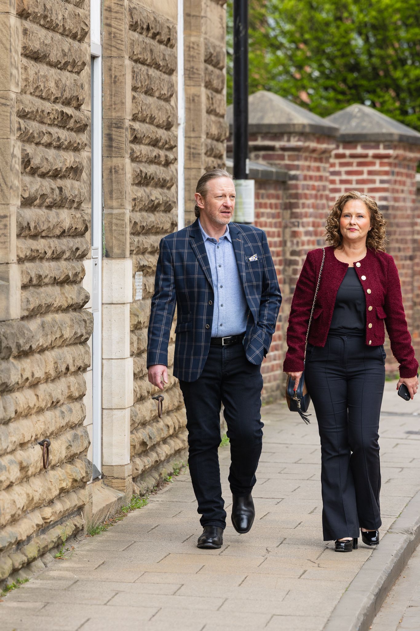 A man and woman in smart casual attire walk together along a brick building sidewalk.