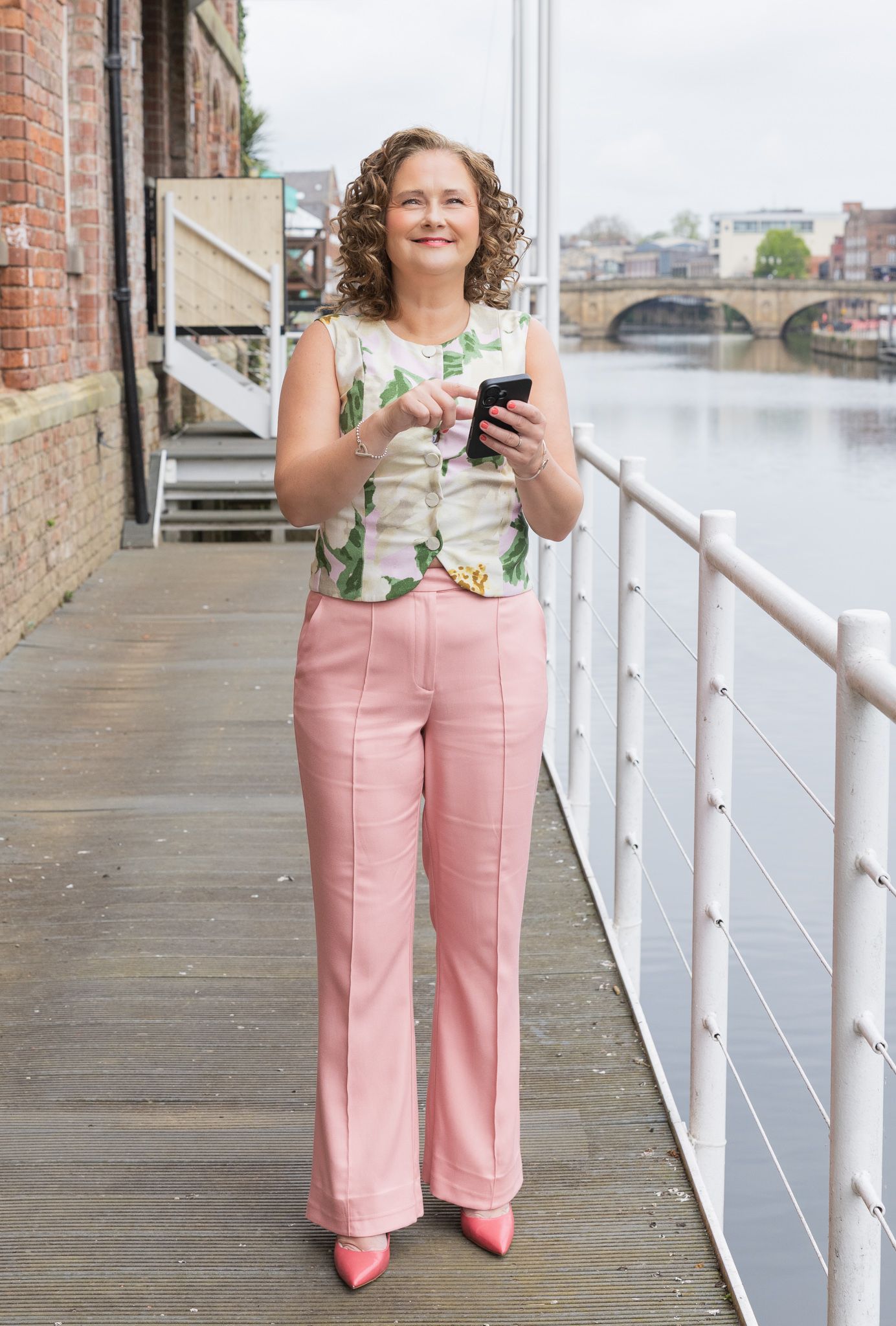 Woman in pink wide-leg trousers and floral top holding phone on waterfront boardwalk near historic bridge.