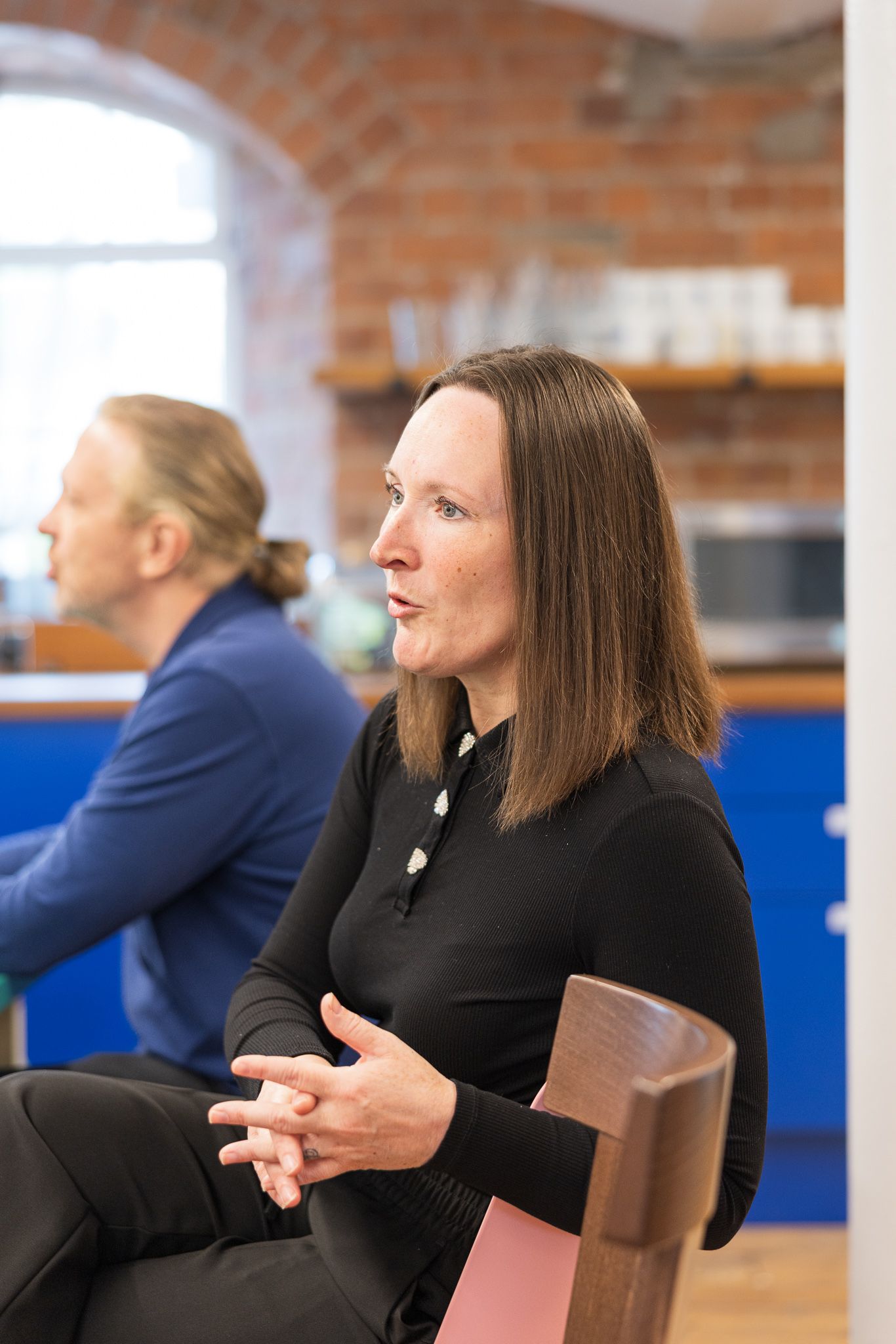 A woman with brown hair in a black turtleneck sits thoughtfully in a modern brick-walled office space.