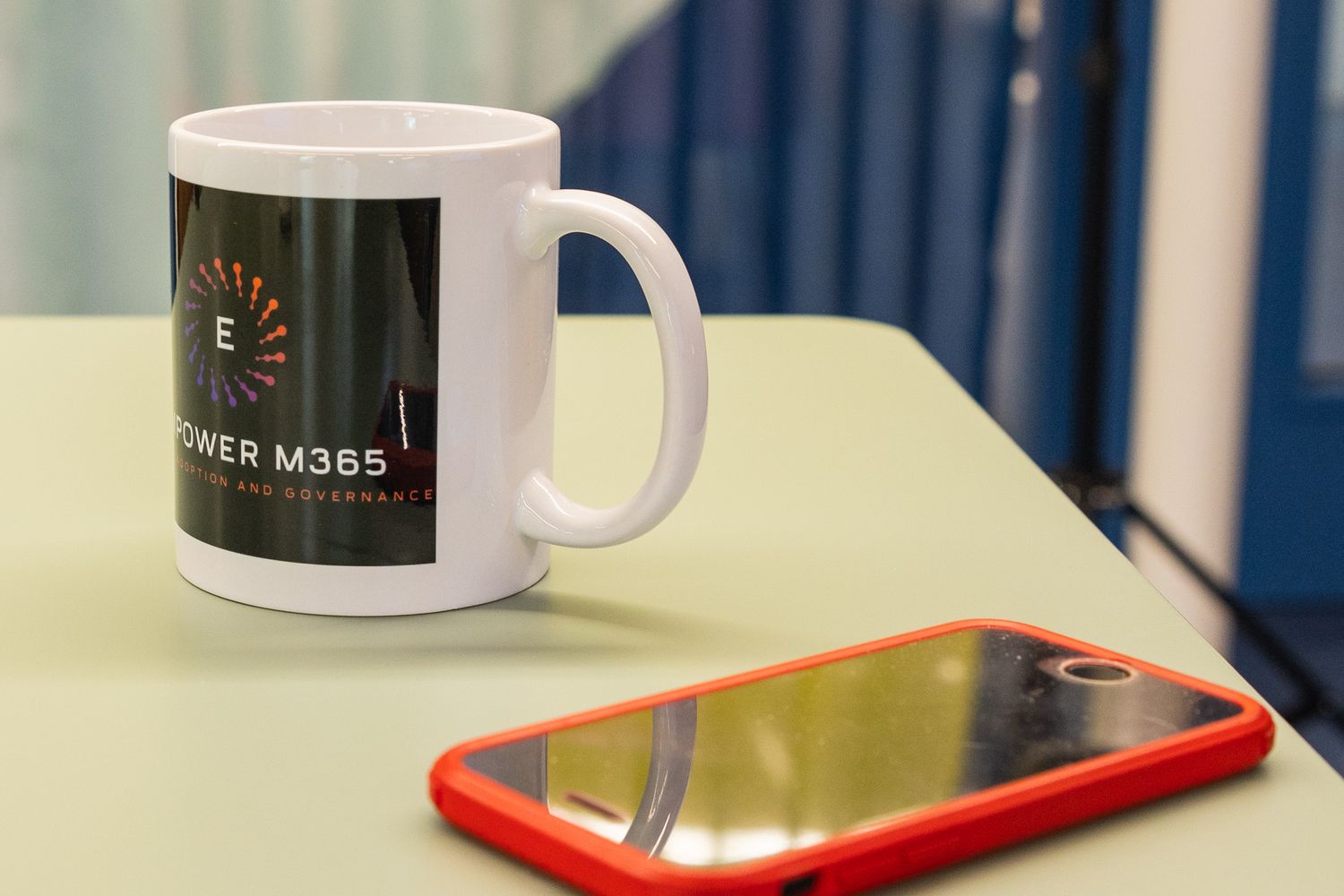 White Power M365 branded coffee mug beside a red-framed smartphone on a light green table.