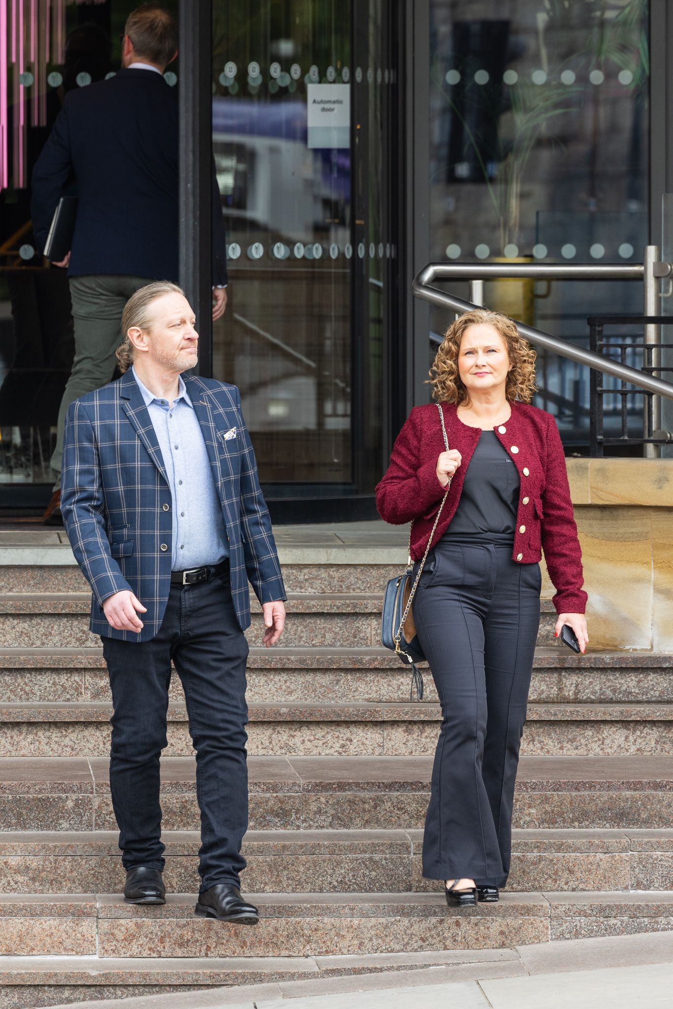 A man in a plaid blazer and woman in a red cardigan walk down stone steps outside a modern building.