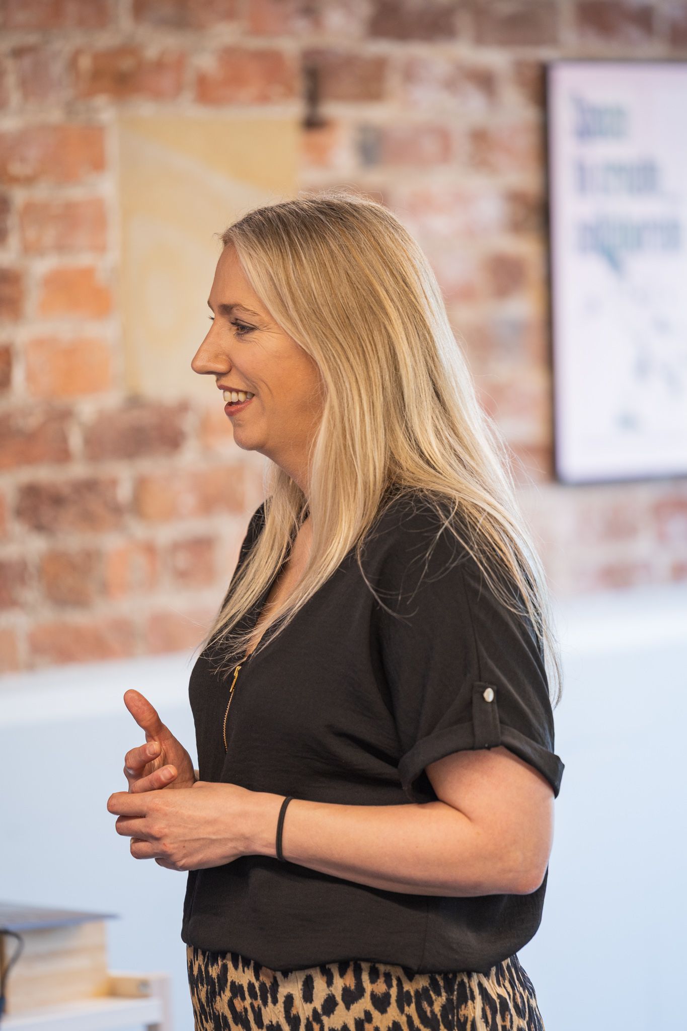 Blonde woman in black top and leopard print skirt speaking at an event in a brick-walled room.