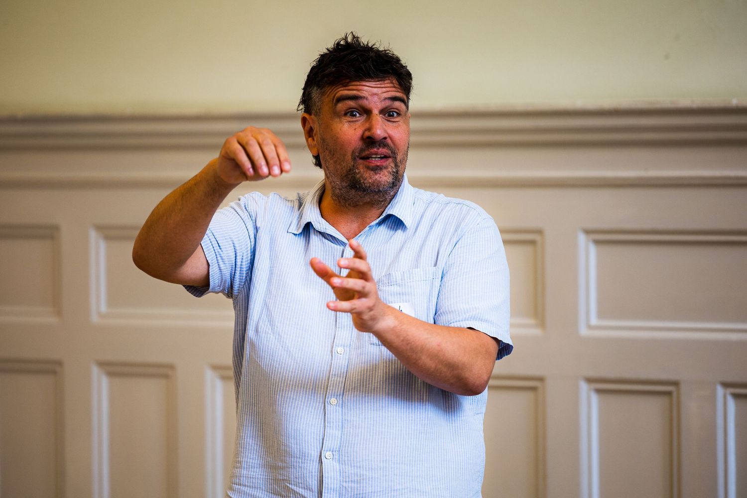 Man in blue shirt gesturing while speaking at an indoor event or presentation.