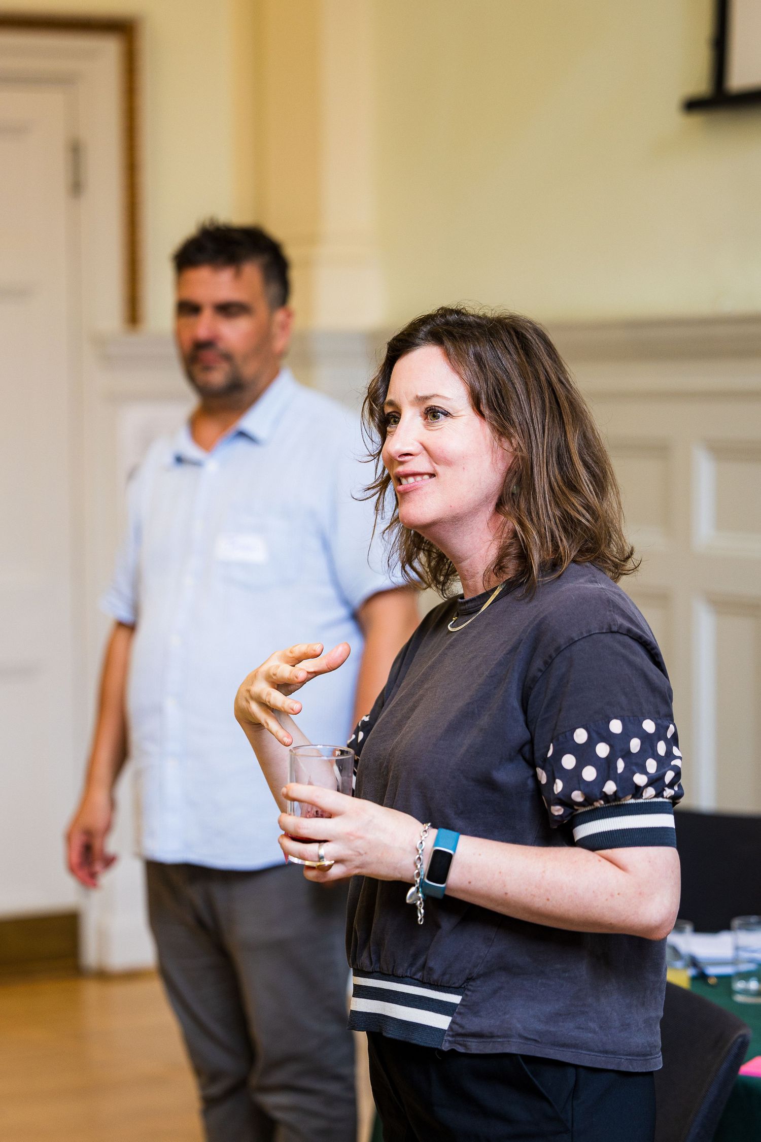 Woman holding a glass drink at an indoor networking event, with a man in a blue shirt standing in the background.