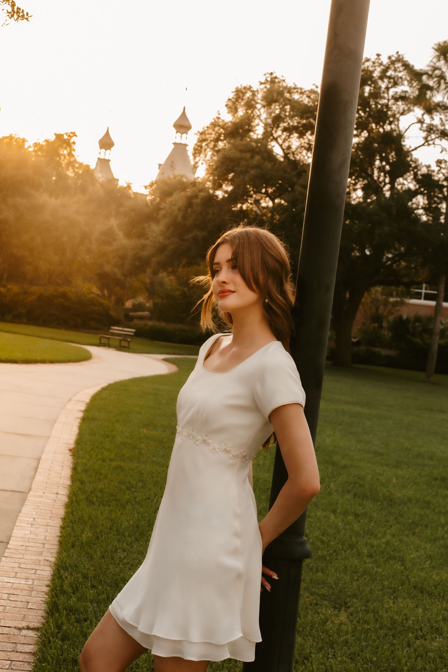 Elegant woman in white mini dress stands by black lamp post at golden hour in scenic park setting.