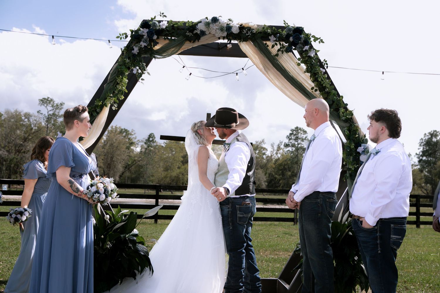 Bride and groom share first kiss under floral arch at outdoor farm wedding with wedding party standing alongside.