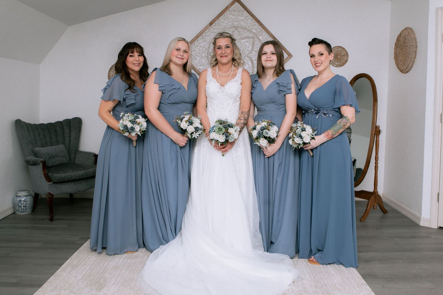 Bride in white gown poses with four bridesmaids in dusty blue dresses holding white bouquets indoors.