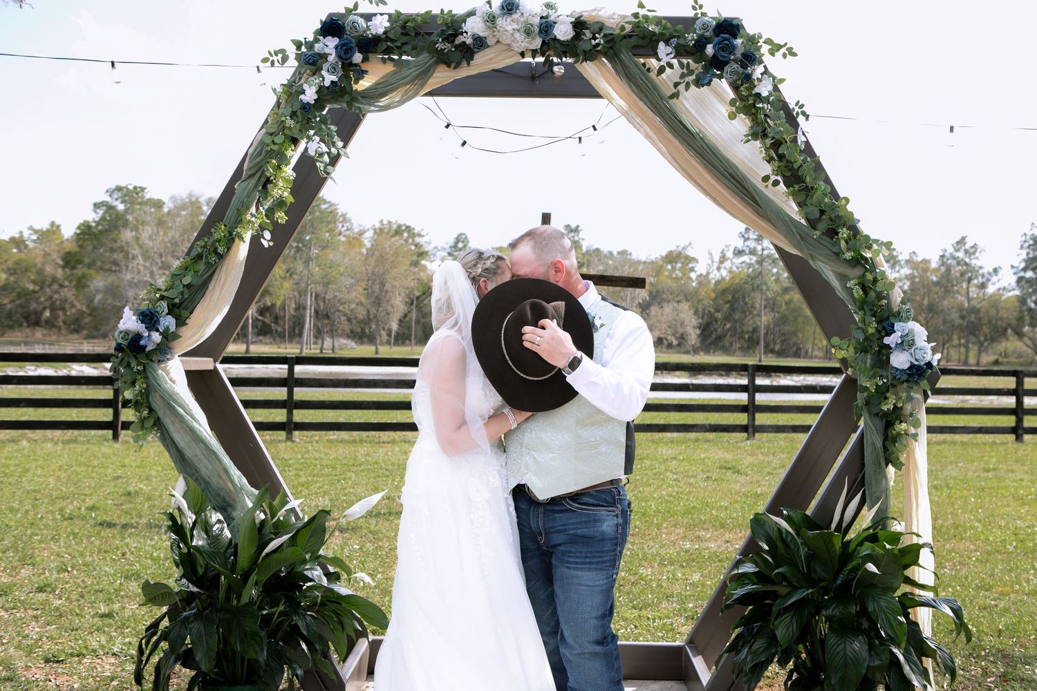 Bride and groom share first kiss under hexagonal wooden arch with greenery at outdoor farm wedding.