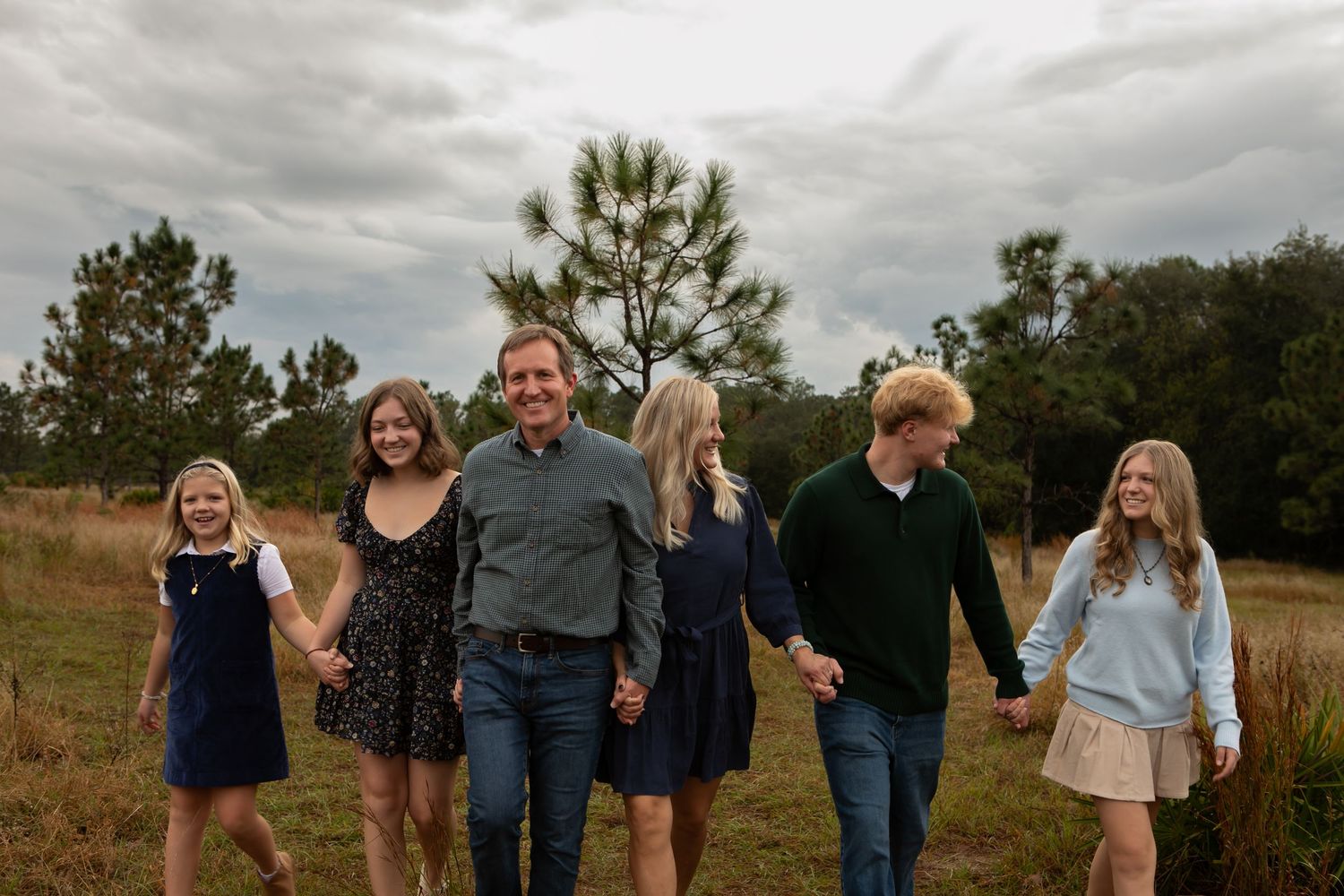 A family of six walks hand in hand through an open grassy field under a cloudy sky.