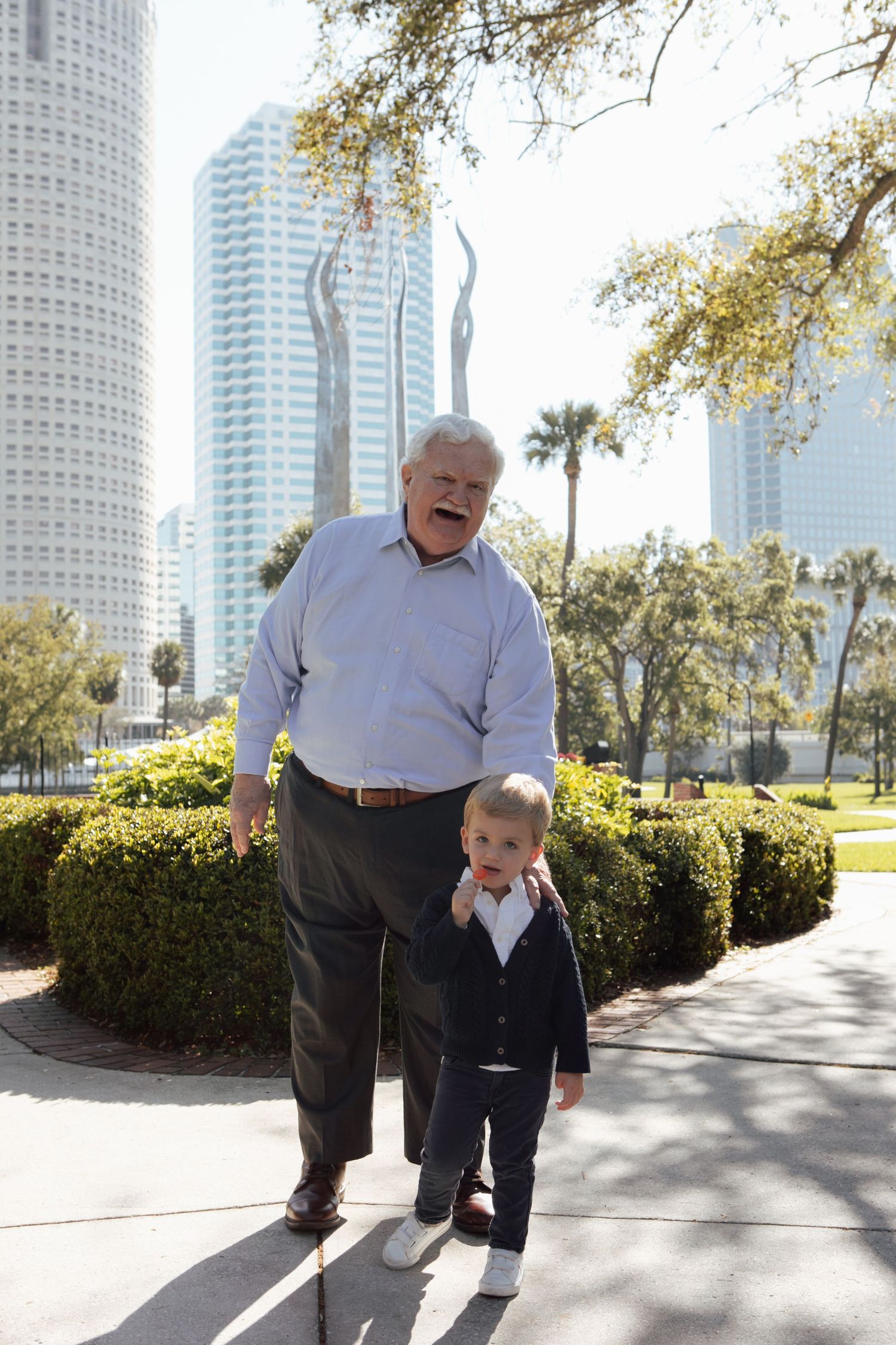 Laughing grandfather walks with dressed-up toddler grandson in front of Tampa urban park skyline.
