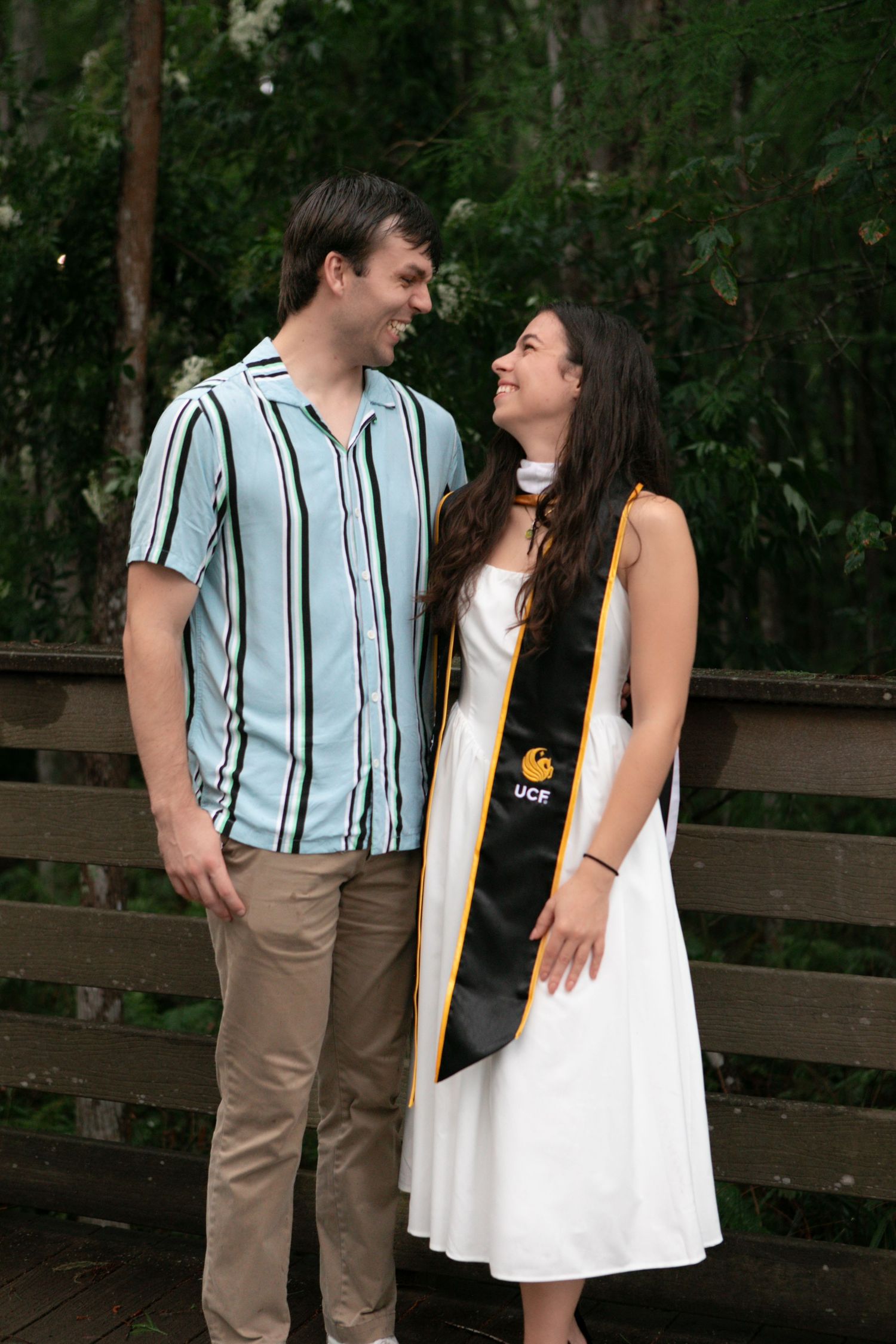 Smiling couple standing together on a wooden bridge, woman wearing a UCF graduation stole.