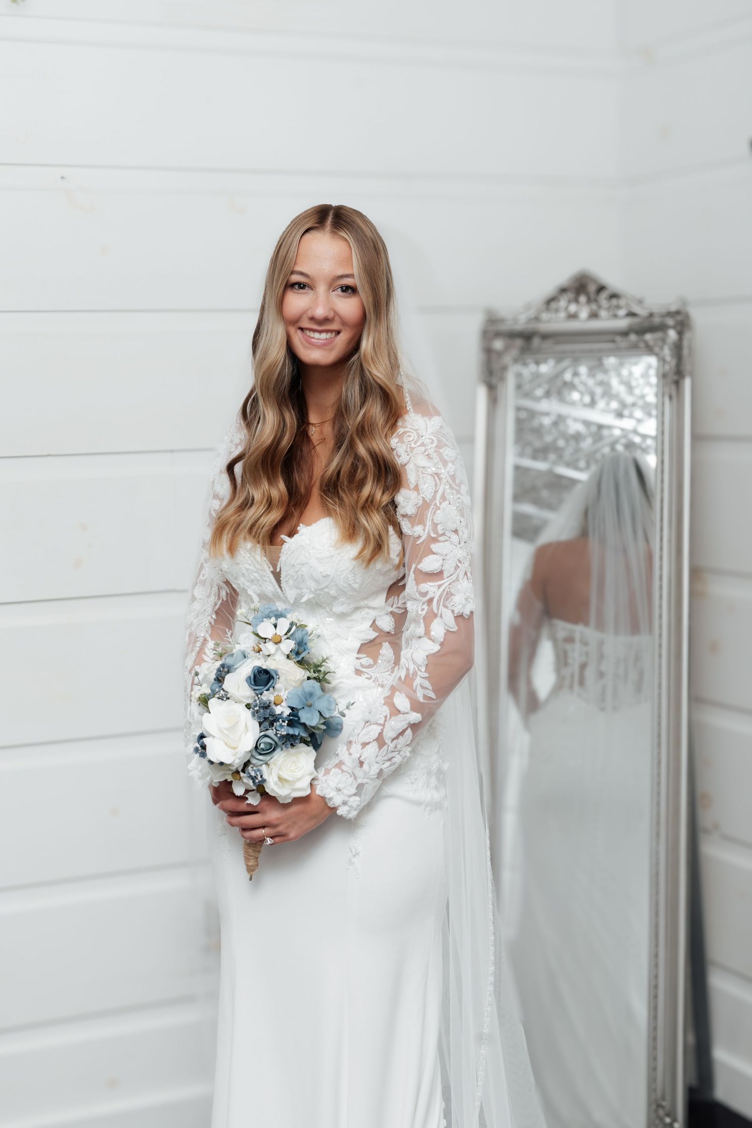 Beautiful smiling bride with long wavy hair holds a blue and white bouquet in a white lace wedding gown.
