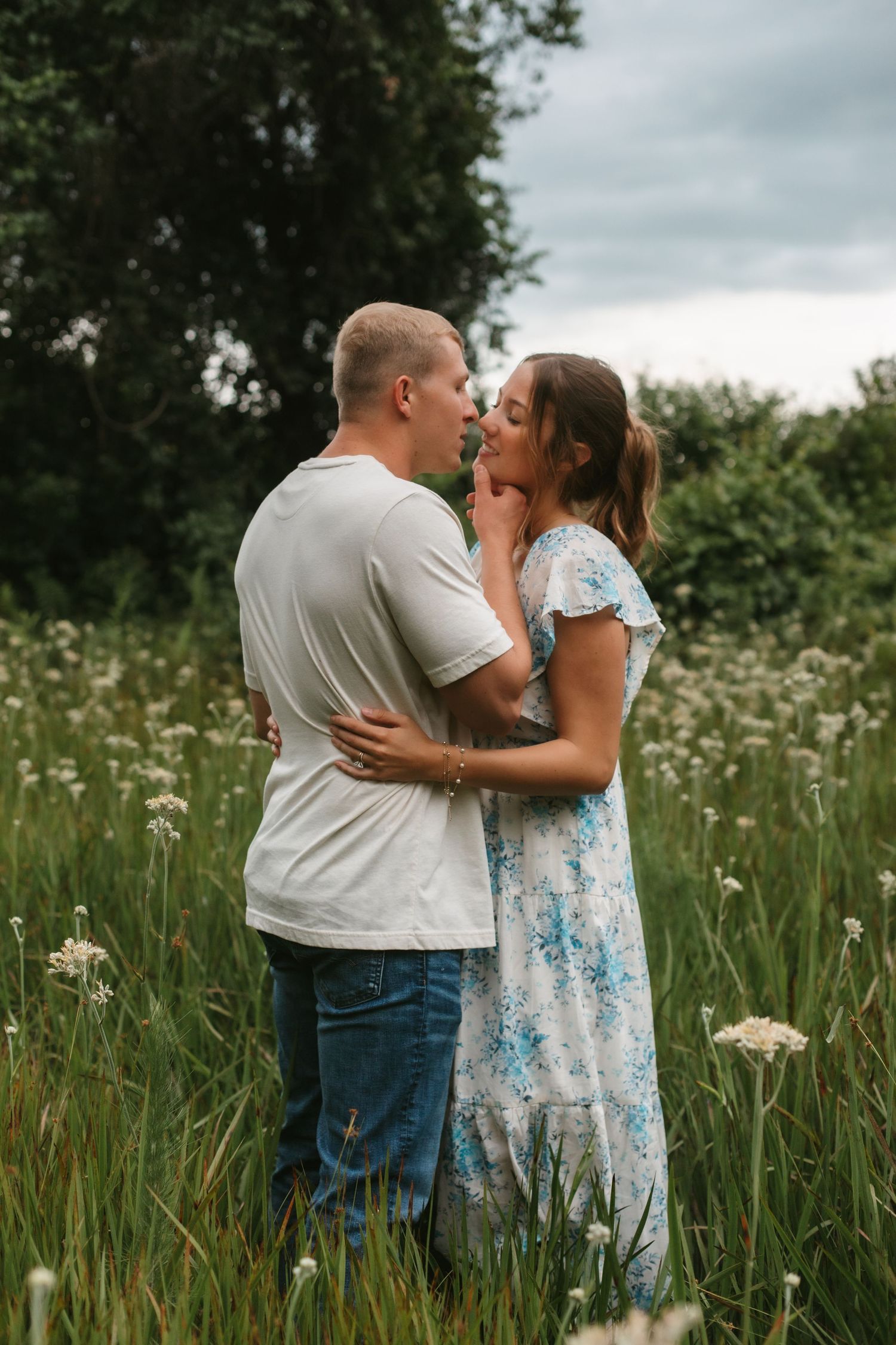 Couple sharing a romantic moment in a lush green meadow with white wildflowers, woman in floral dress embracing man in white tee.