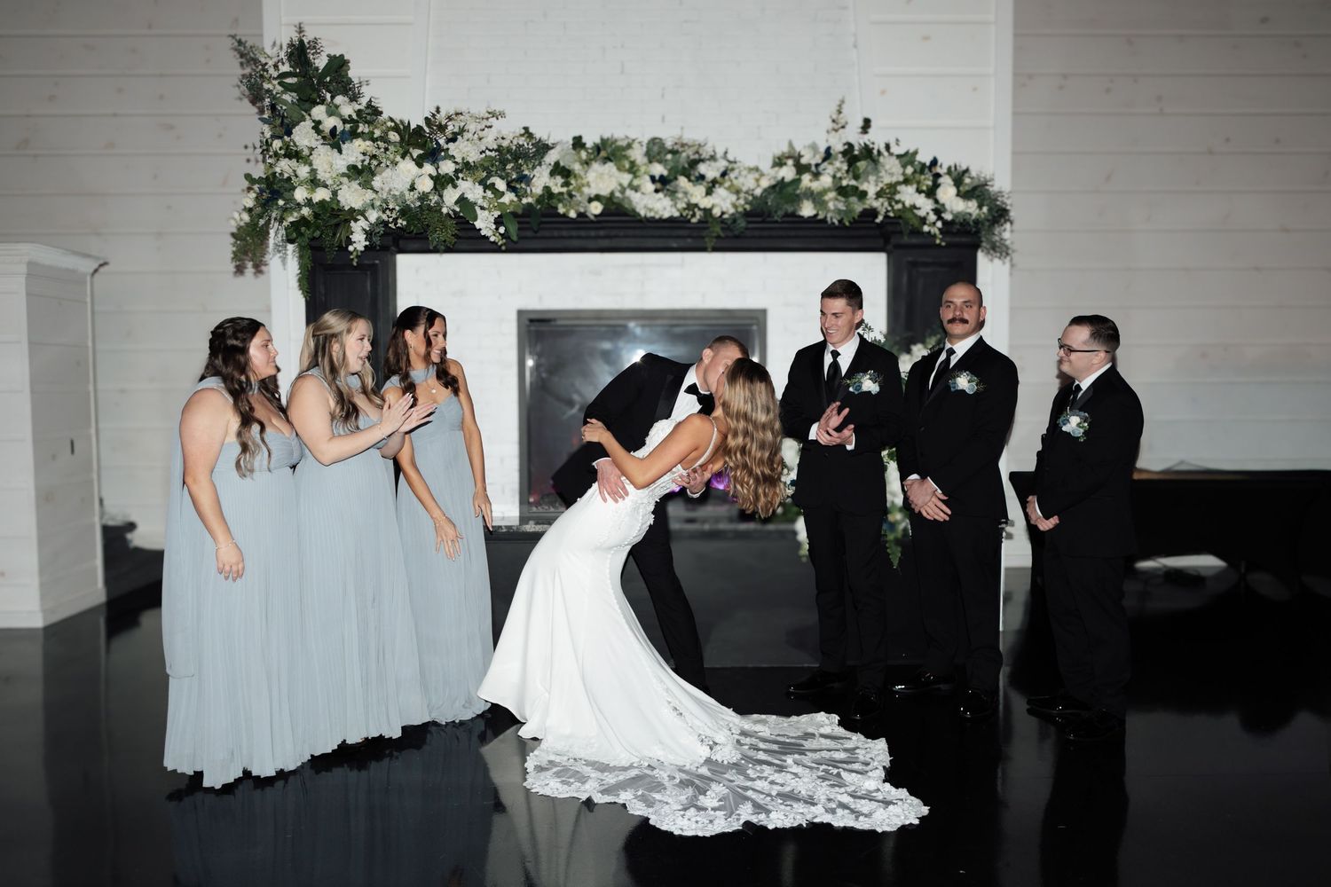 Bride and groom share a dip kiss surrounded by bridesmaids in dusty blue gowns and groomsmen near a floral fireplace.
