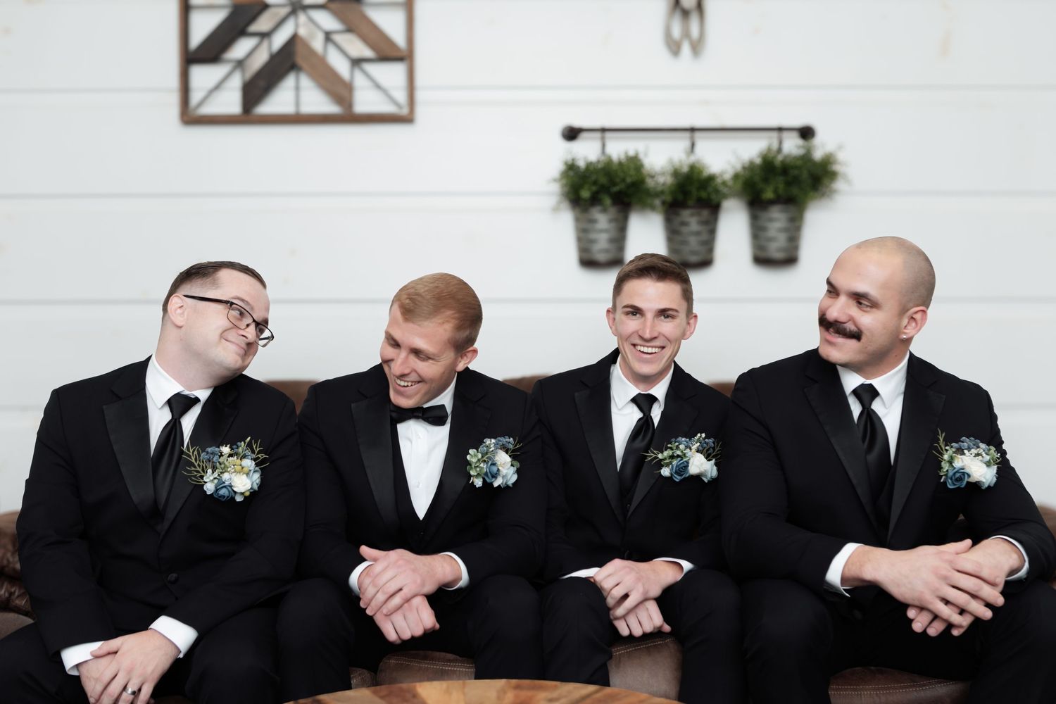 Four groomsmen in black tuxedos with floral boutonnieres sitting on a leather couch laughing at a wedding.