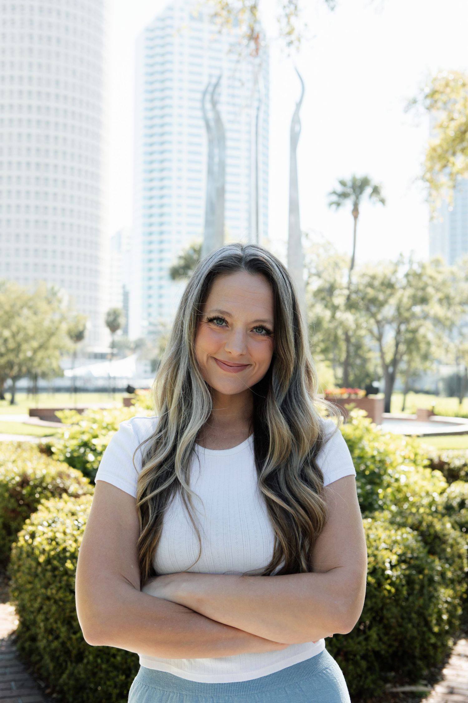 Smiling woman with long blonde highlighted hair poses confidently with arms crossed in front of city skyscrapers.