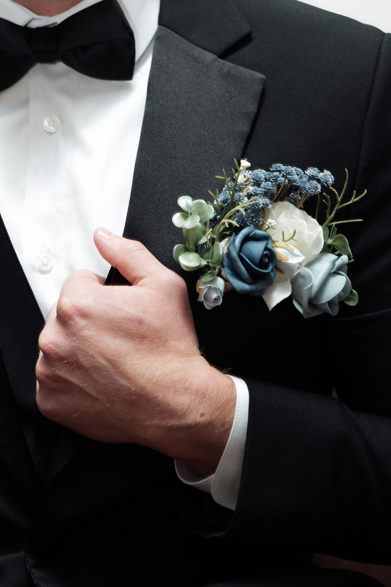 Groom adjusts his blue and white floral boutonniere on his black tuxedo with bow tie.
