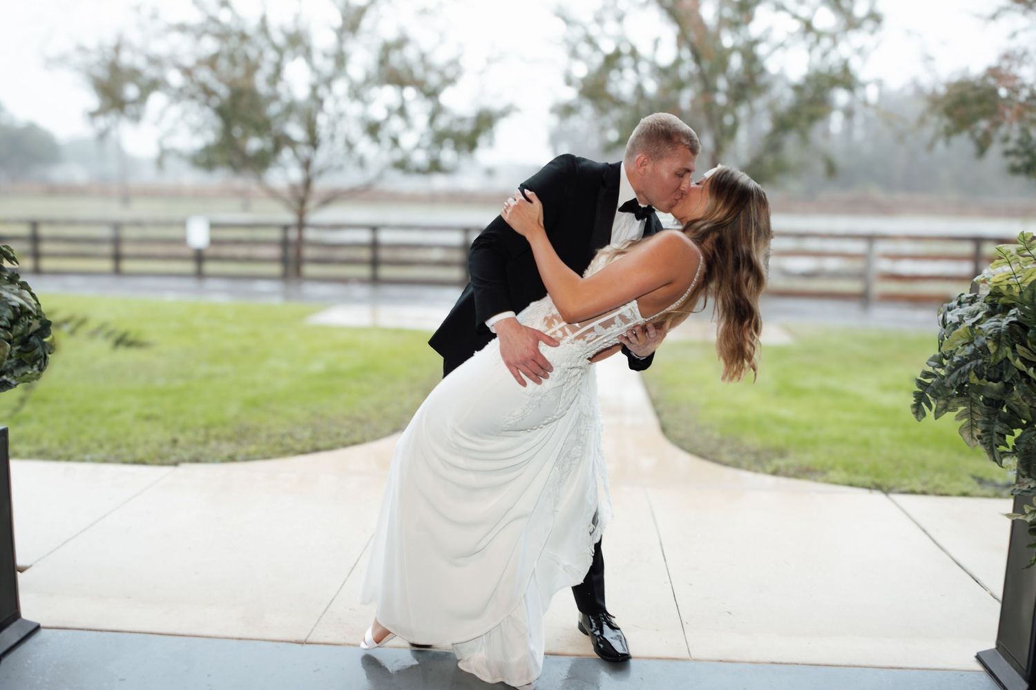 Groom dips bride in elegant white wedding dress outdoors at rustic venue with wooden fence backdrop.