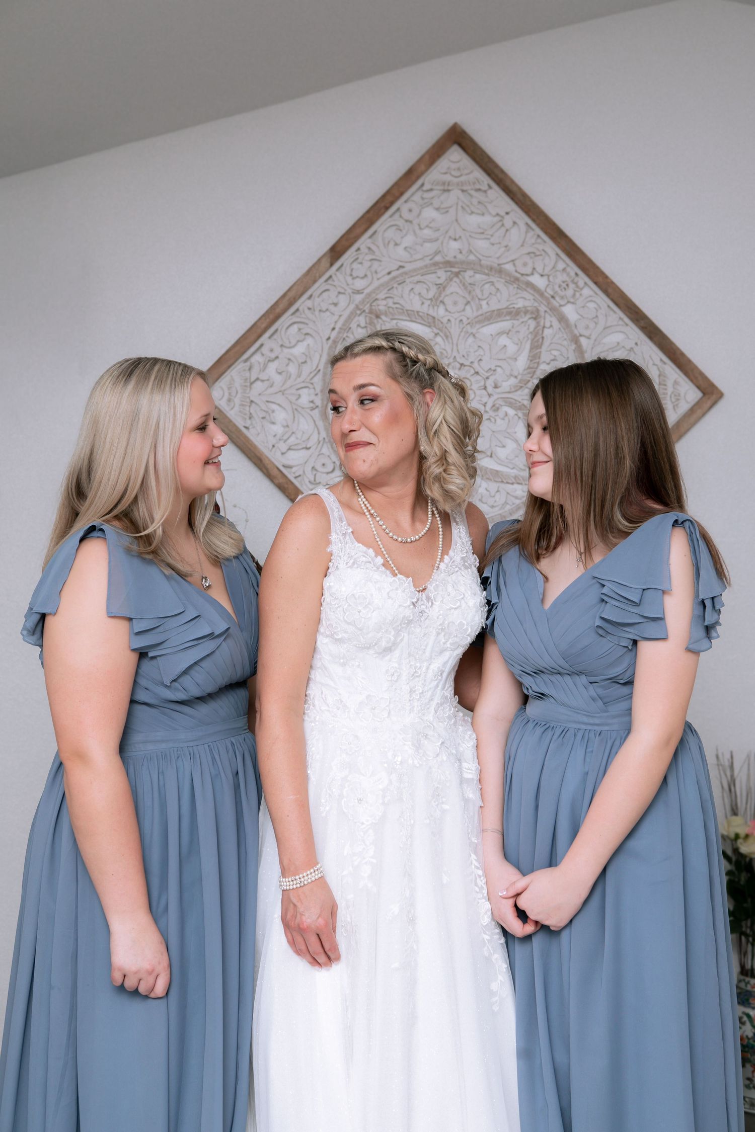 Bride in white lace gown stands between two bridesmaids in dusty blue dresses, posing together before the wedding ceremony.