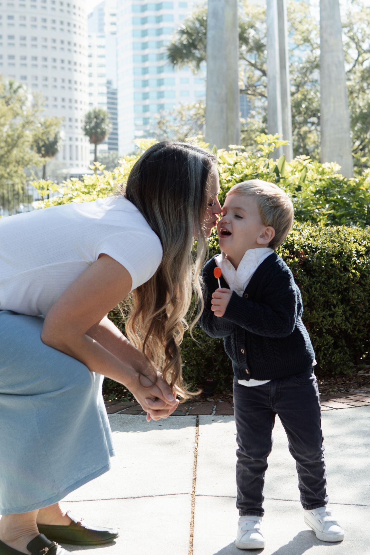 Mother leans in to kiss toddler son holding orange lollipop in sunny urban park setting.