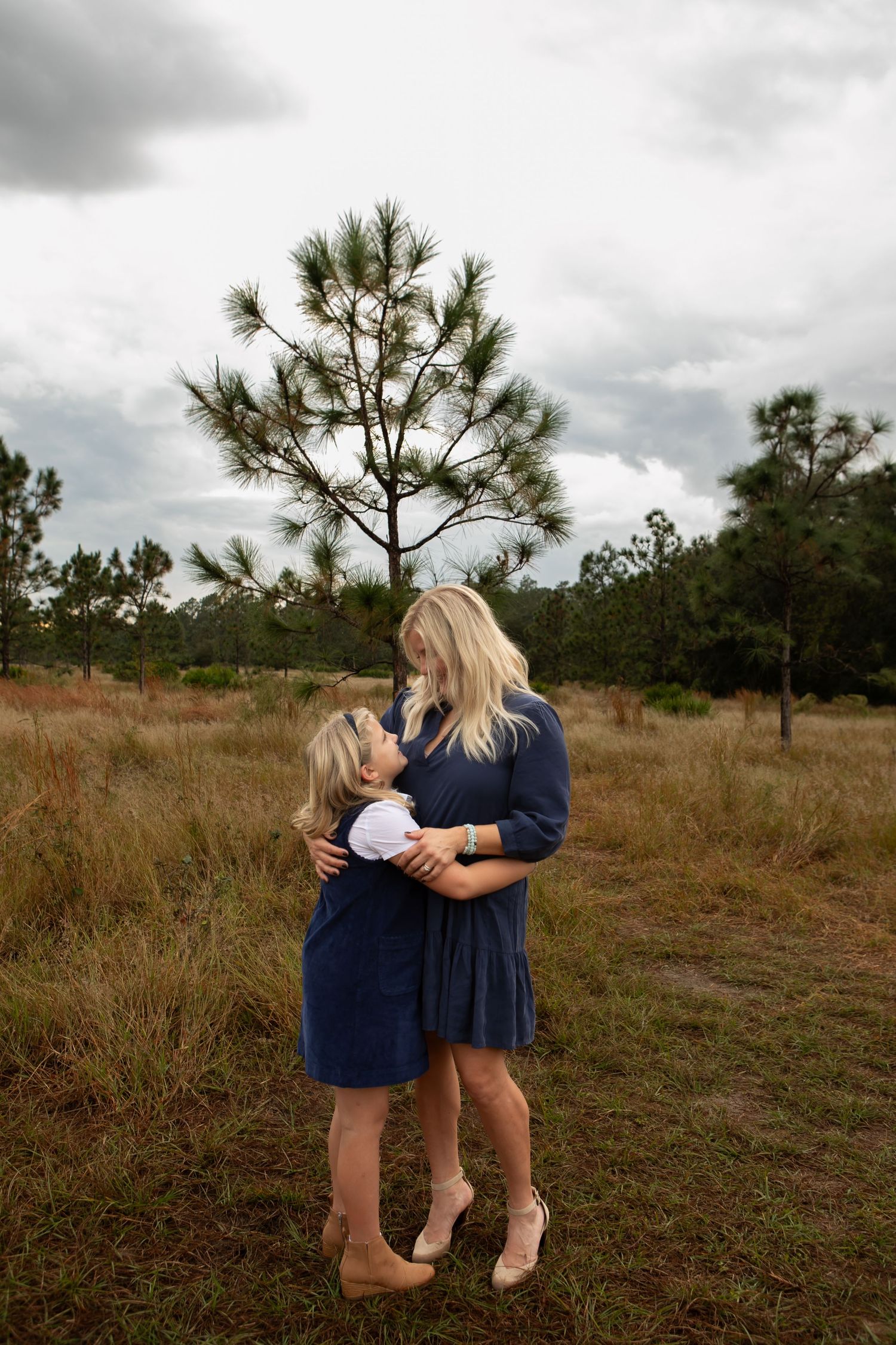 A blonde woman in a navy dress tenderly embraces a young girl in a grassy outdoor field.