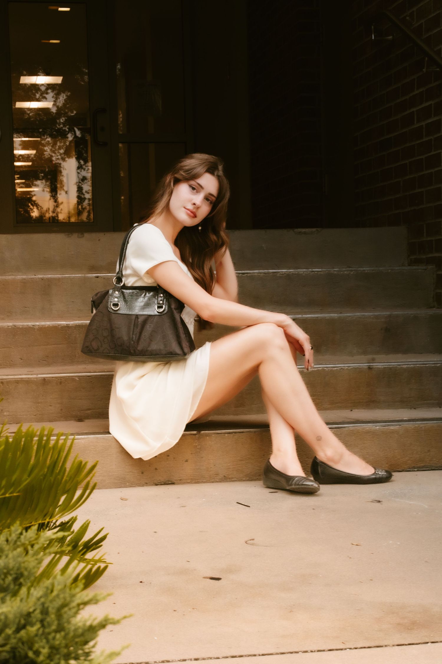 Young woman in white dress with black handbag sitting on steps outside a brick building.