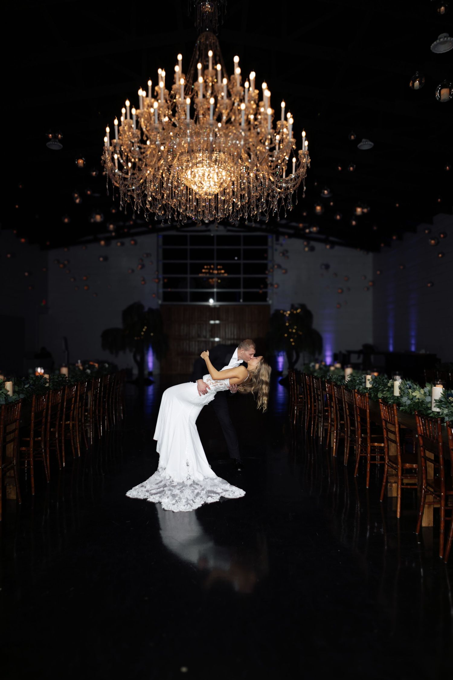 Bride and groom share romantic dip kiss under grand chandelier in elegant reception hall.