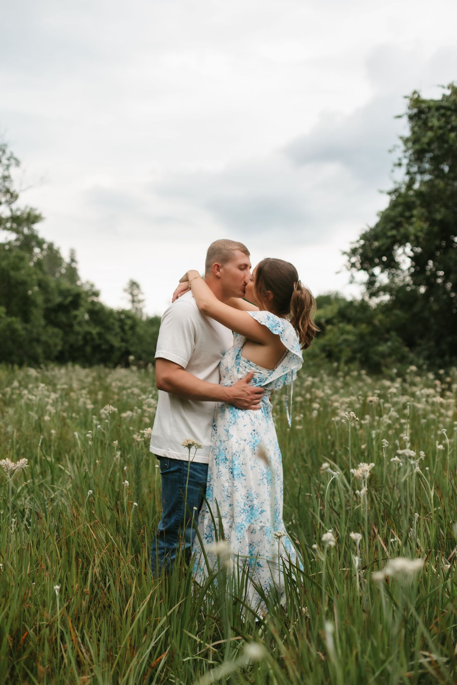 Couple kissing in a lush wildflower meadow, woman in blue floral dress, romantic engagement photo outdoors.