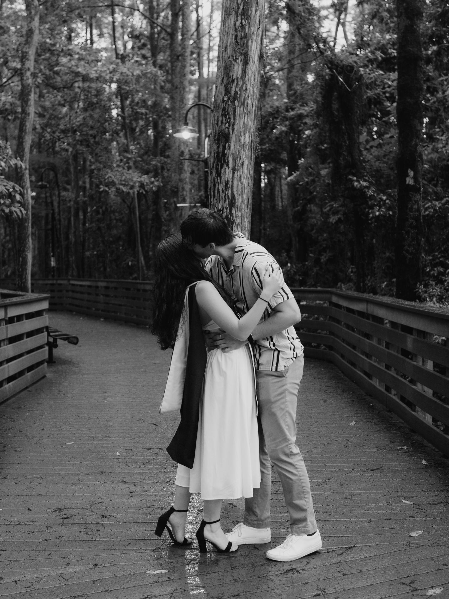 Black and white photo of a couple embracing and kissing on a wooden bridge in a forest.