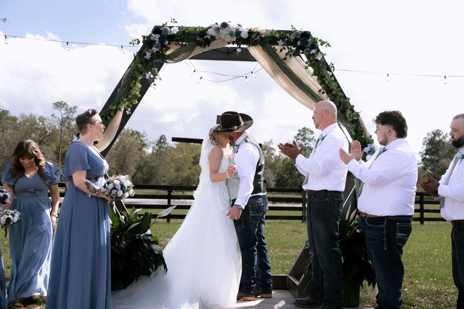 Bride and groom share first kiss under floral arch at outdoor wedding ceremony with bridal party.