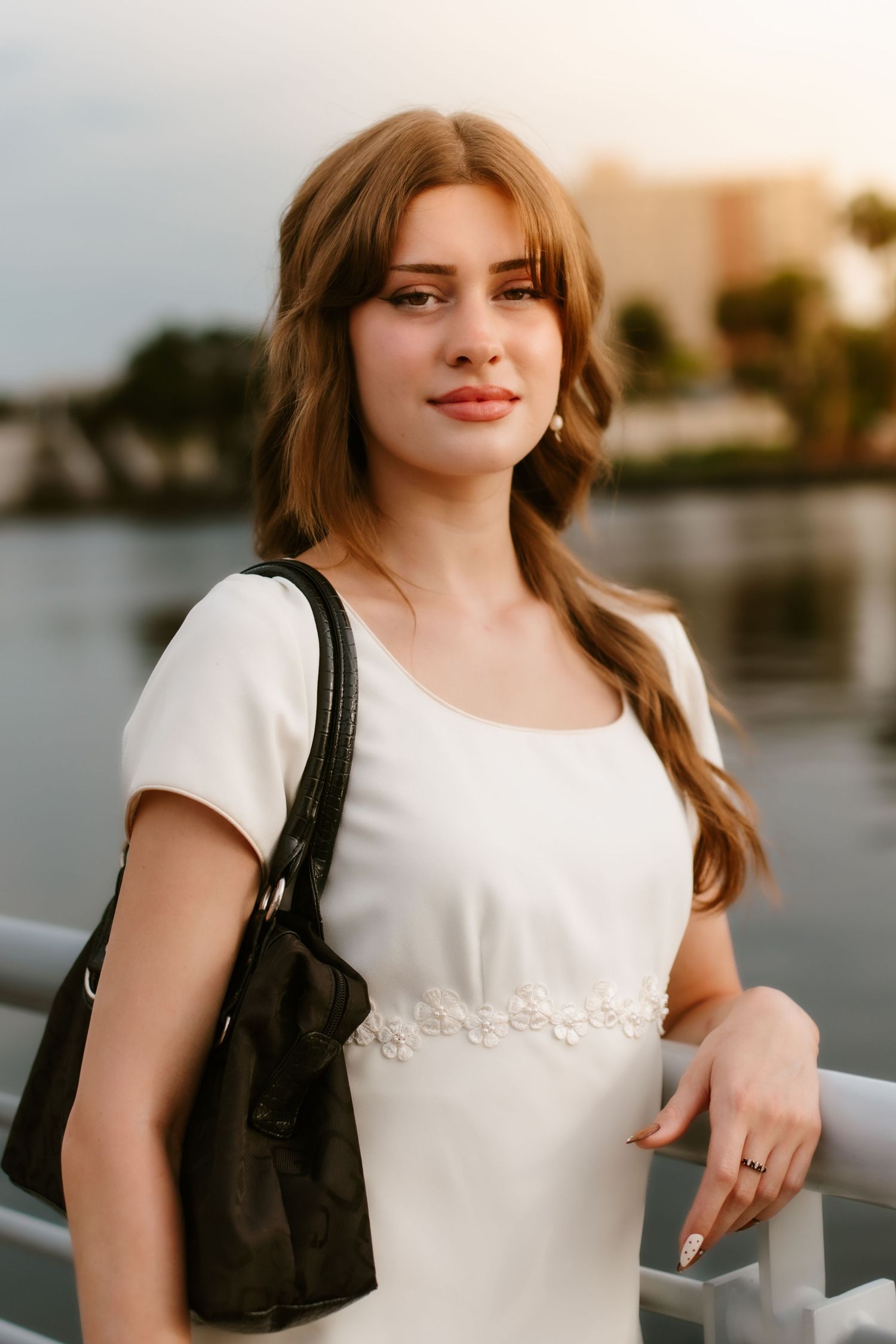 Young woman in white dress with floral detail, carrying black bag, posing by waterfront railing at sunset.