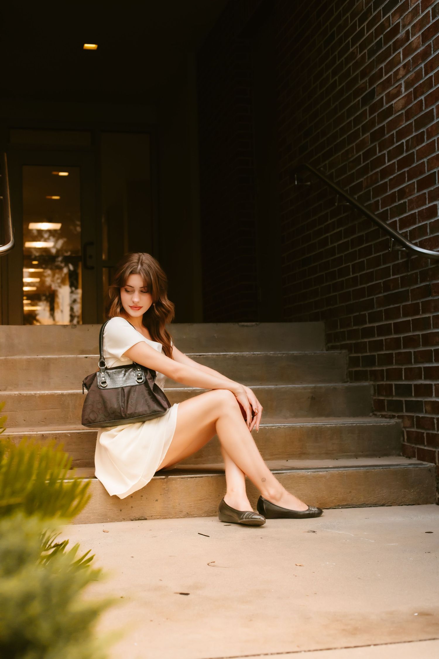 Young woman in white dress and flats sits on brick building steps with dark handbag, posed elegantly outdoors.