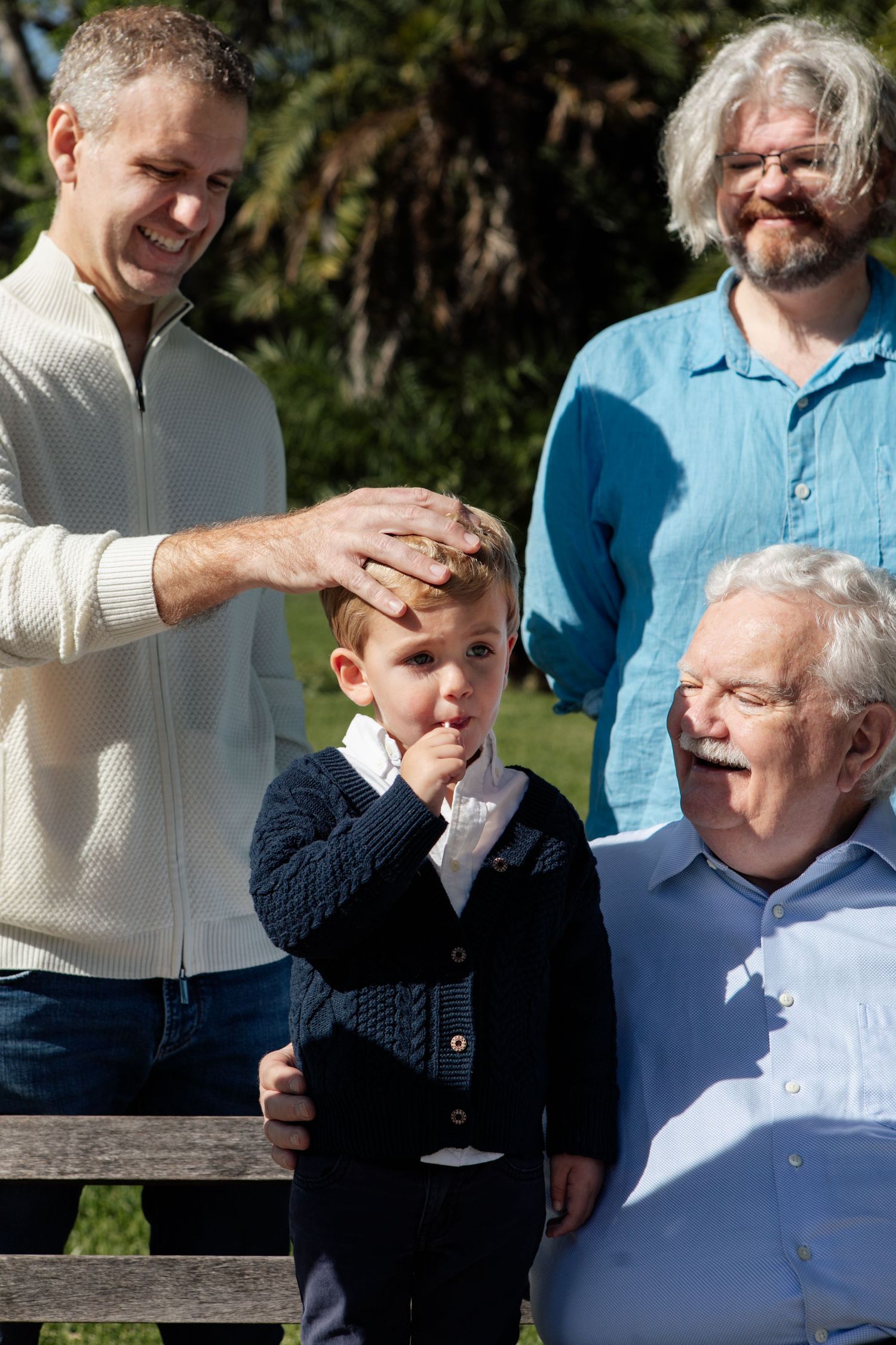 Three generations of men and a young boy pose together outdoors in a sunny park setting.