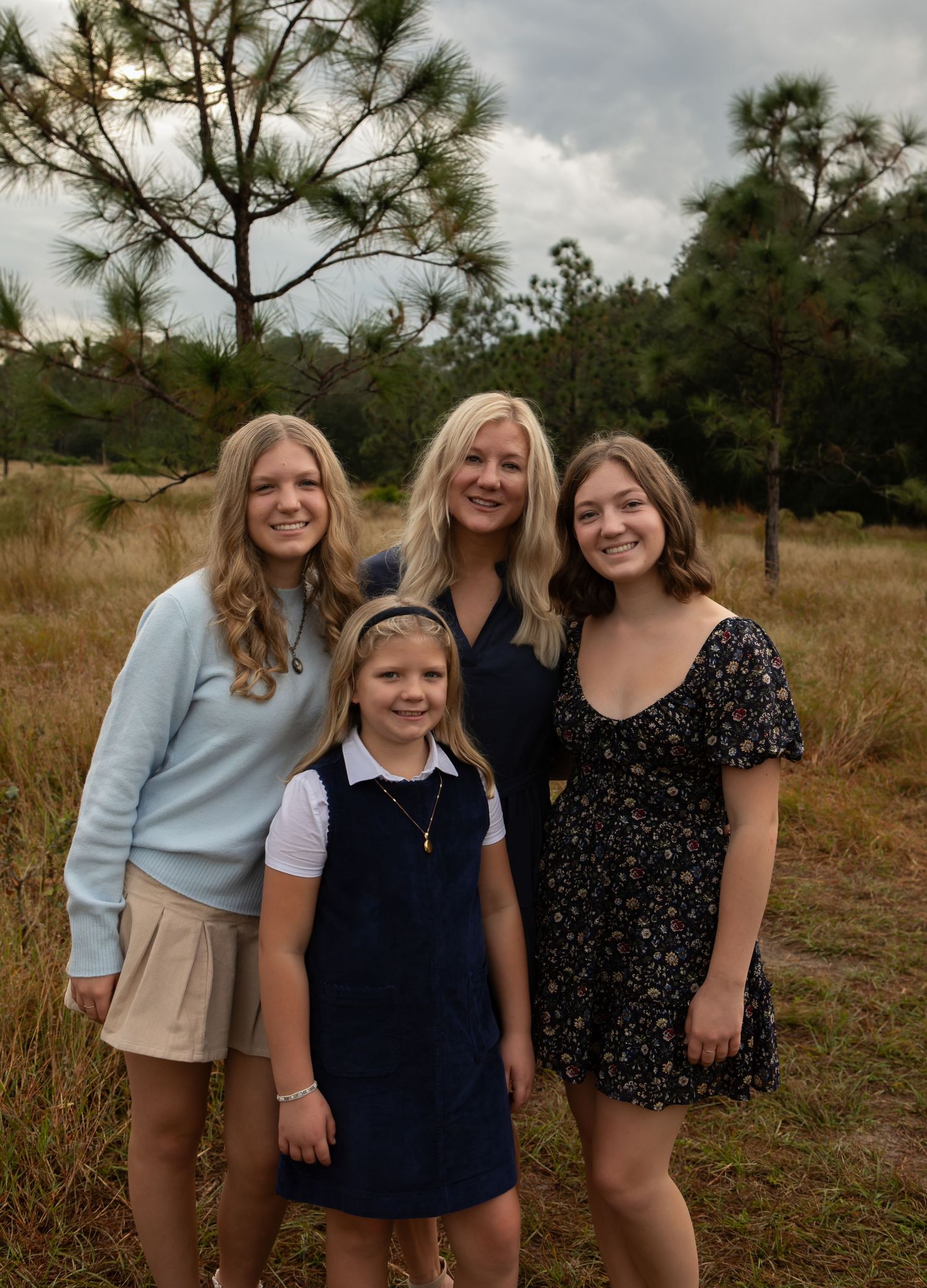 A woman and three girls pose together smiling outdoors in a grassy field with pine trees under a cloudy sky.