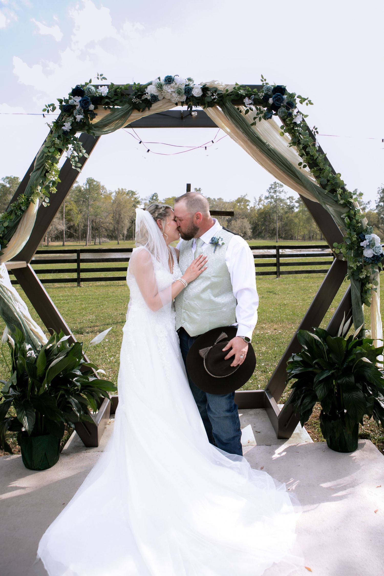 Bride and groom share first kiss under hexagonal floral arch decorated with blue and white flowers at farm wedding.