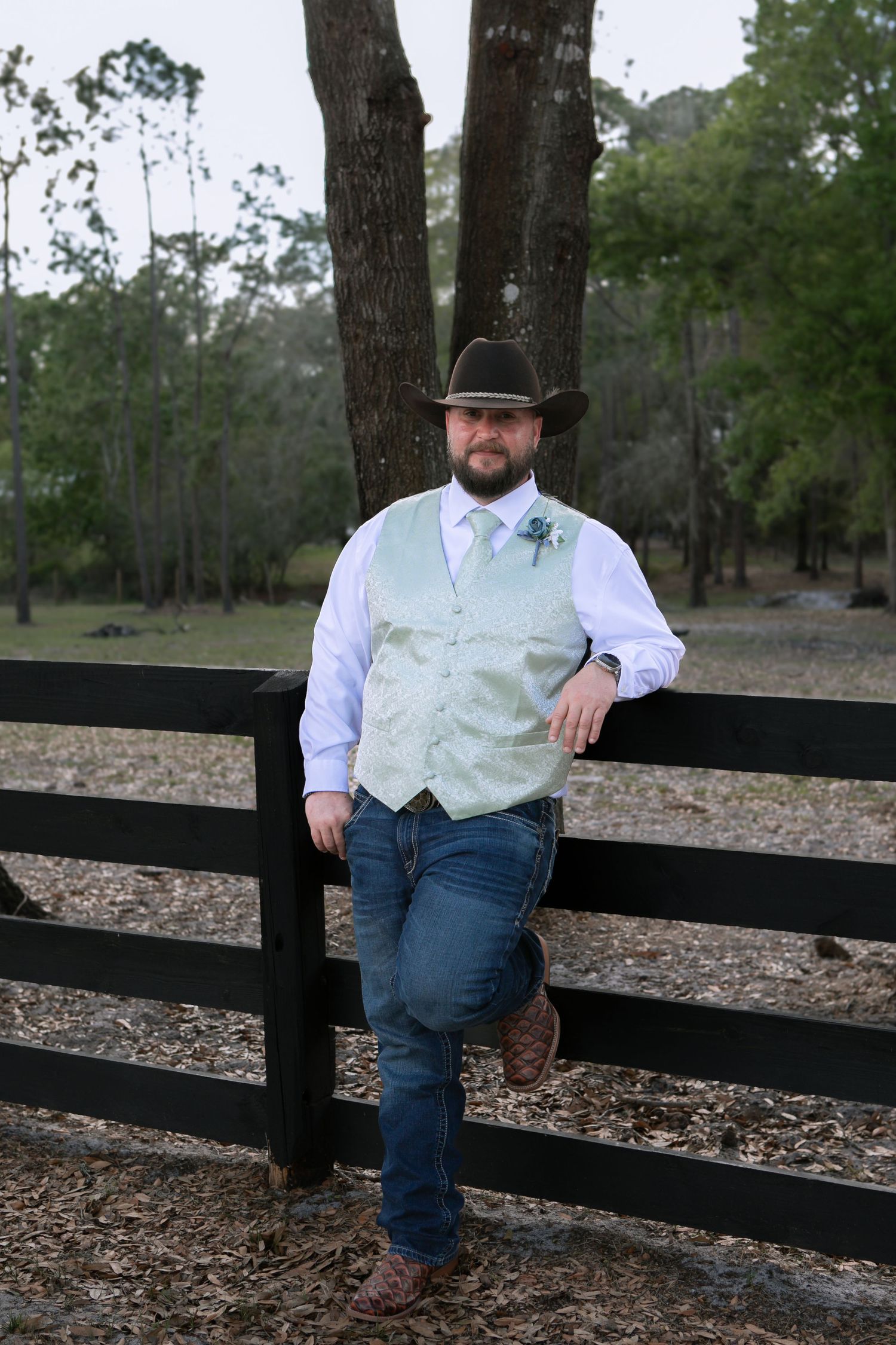 Groom in cowboy hat, mint vest, jeans, and boots leaning against black fence at outdoor rustic wedding venue.