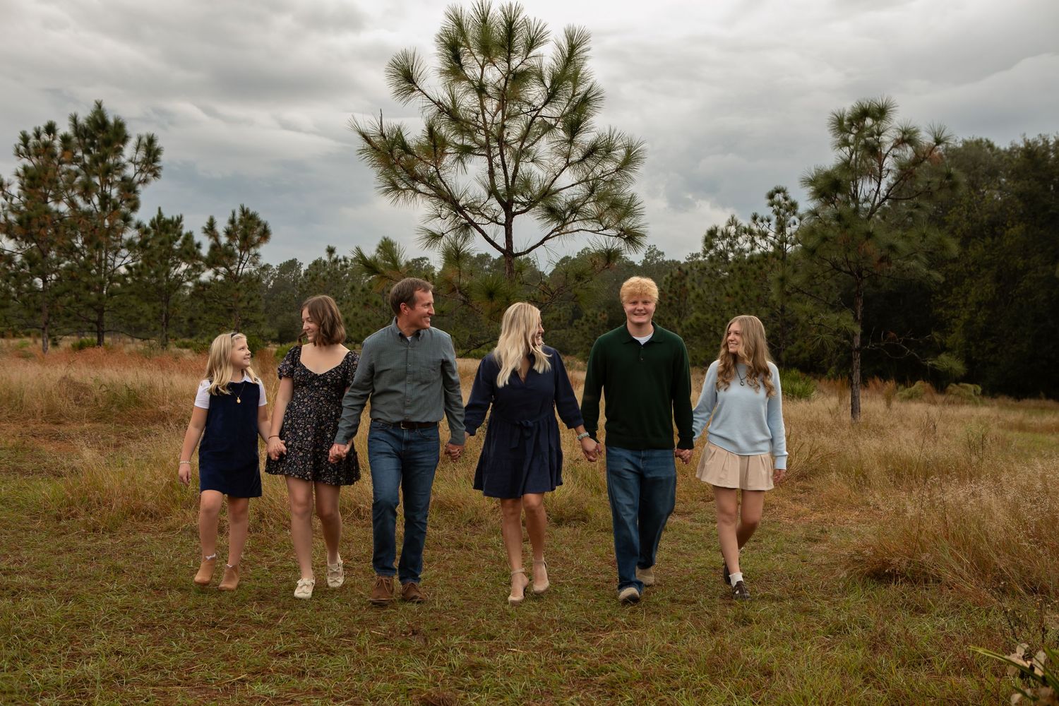 Family of five walking hand-in-hand through an open grassy field with pine trees.