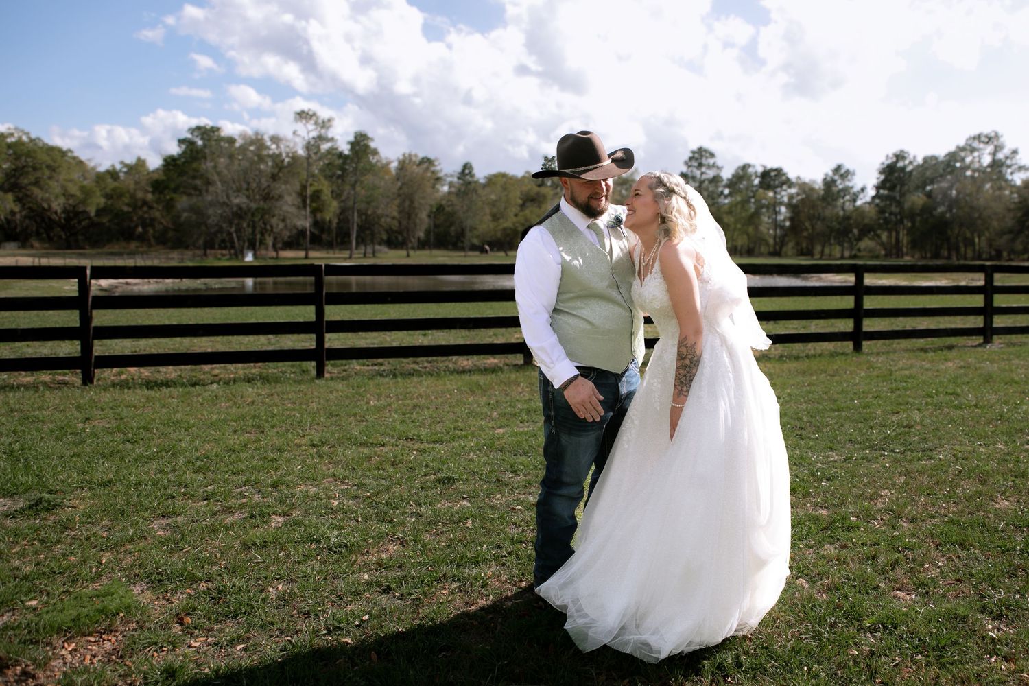 Bride and groom share a romantic moment in a rustic farm setting with wooden fences and green pastures on their wedding day.