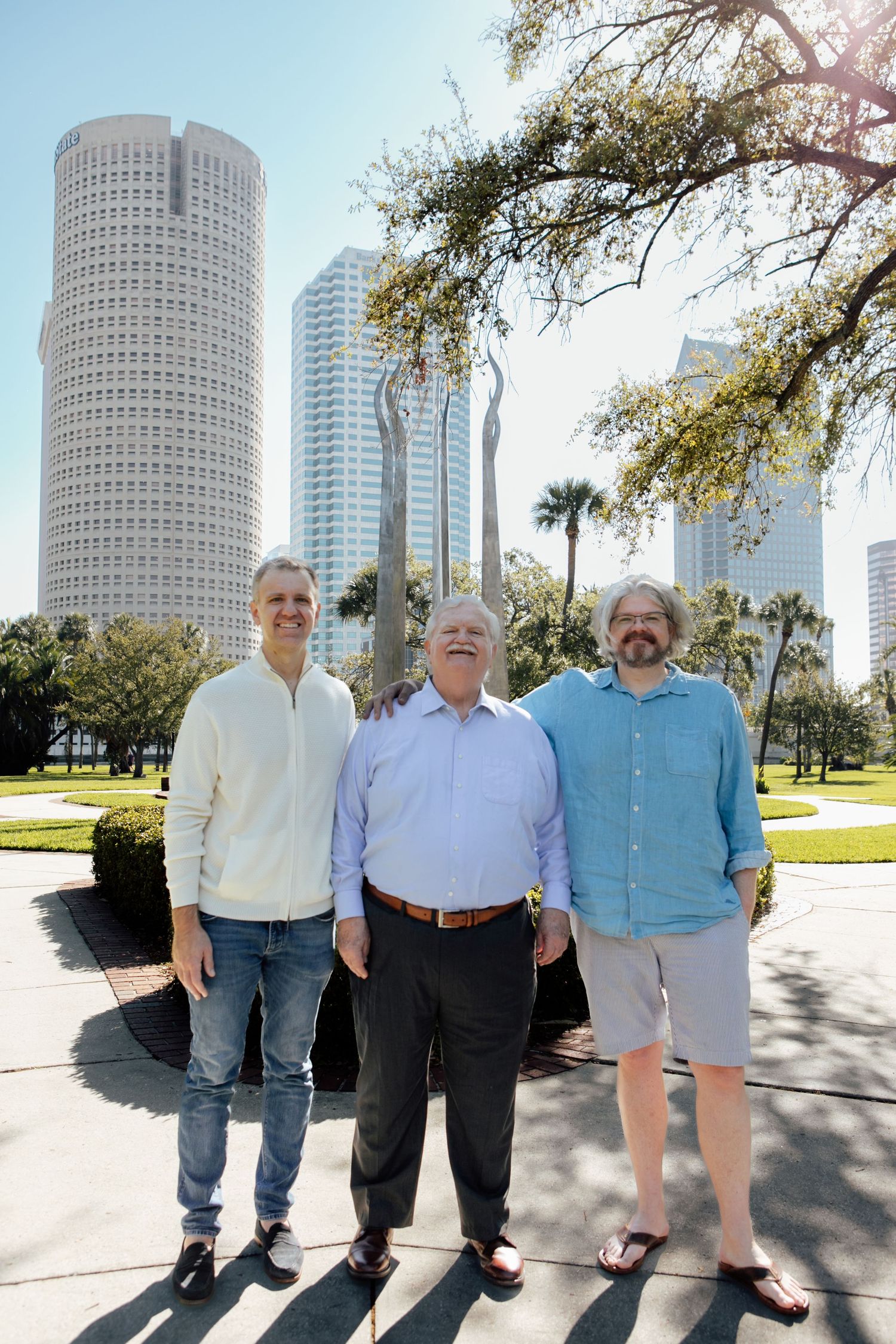 Three men smile together in front of a city skyline with tall buildings and palm trees.