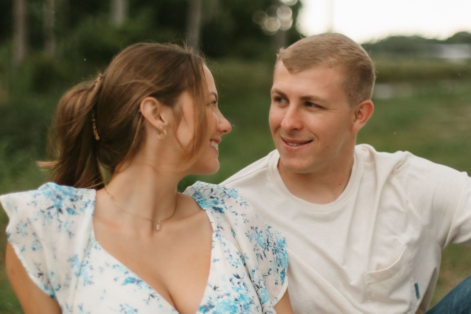 A smiling couple gazes at each other outdoors, woman in floral dress and man in white t-shirt.