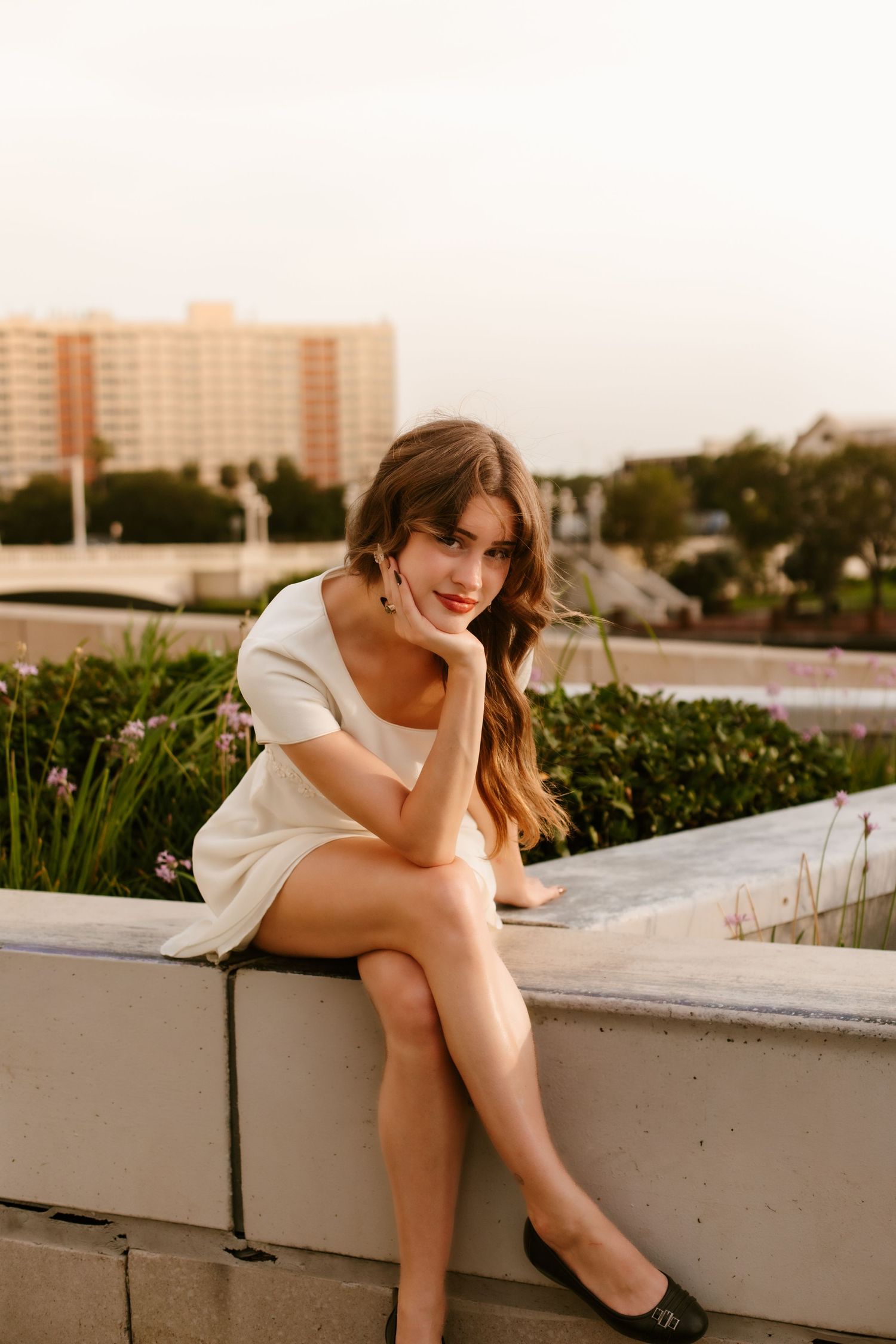 Young woman in white mini dress sitting on a concrete ledge outdoors, smiling with city buildings in background.