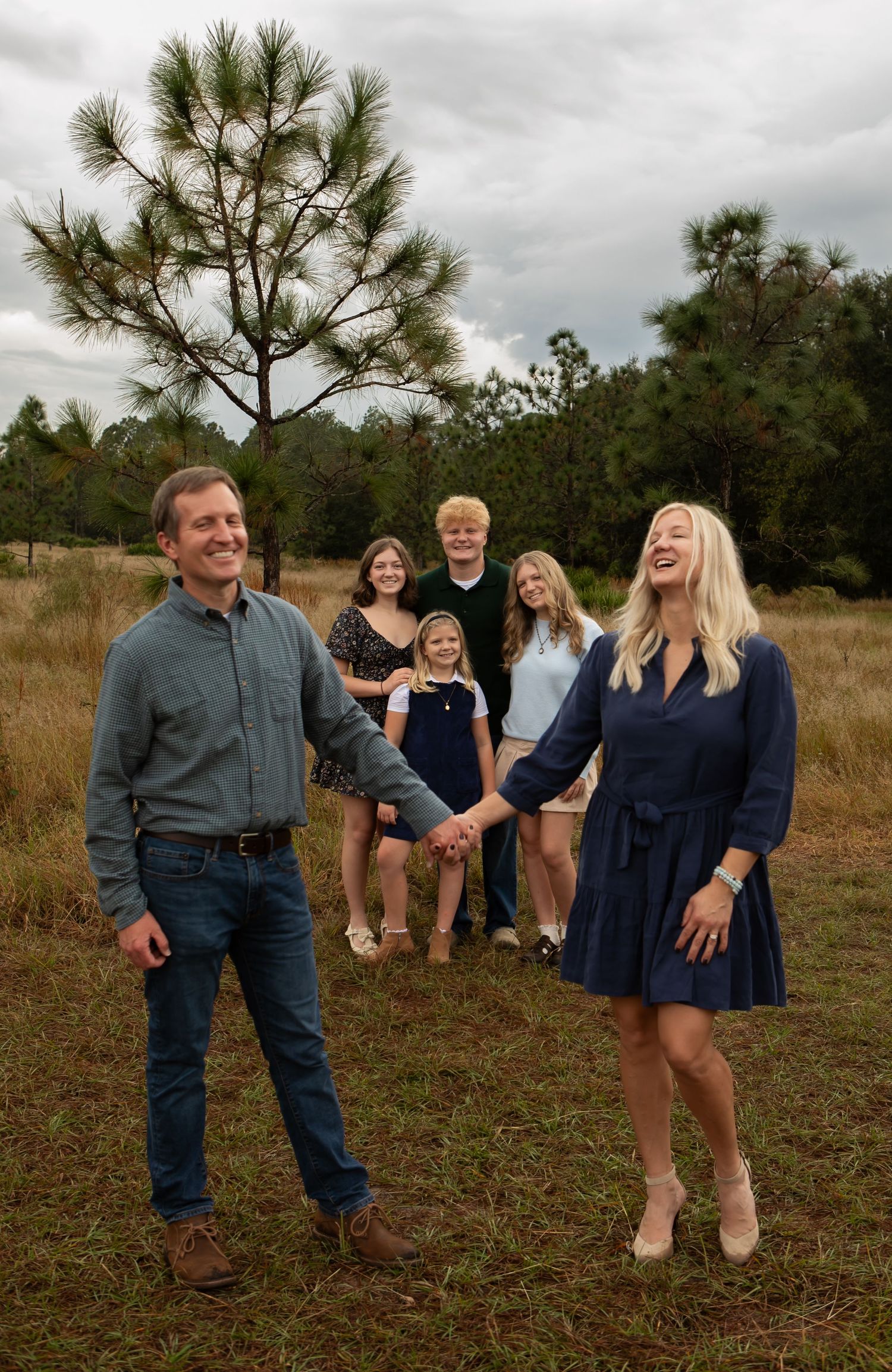 Happy family of six holding hands in a rustic field with pine trees under cloudy skies.