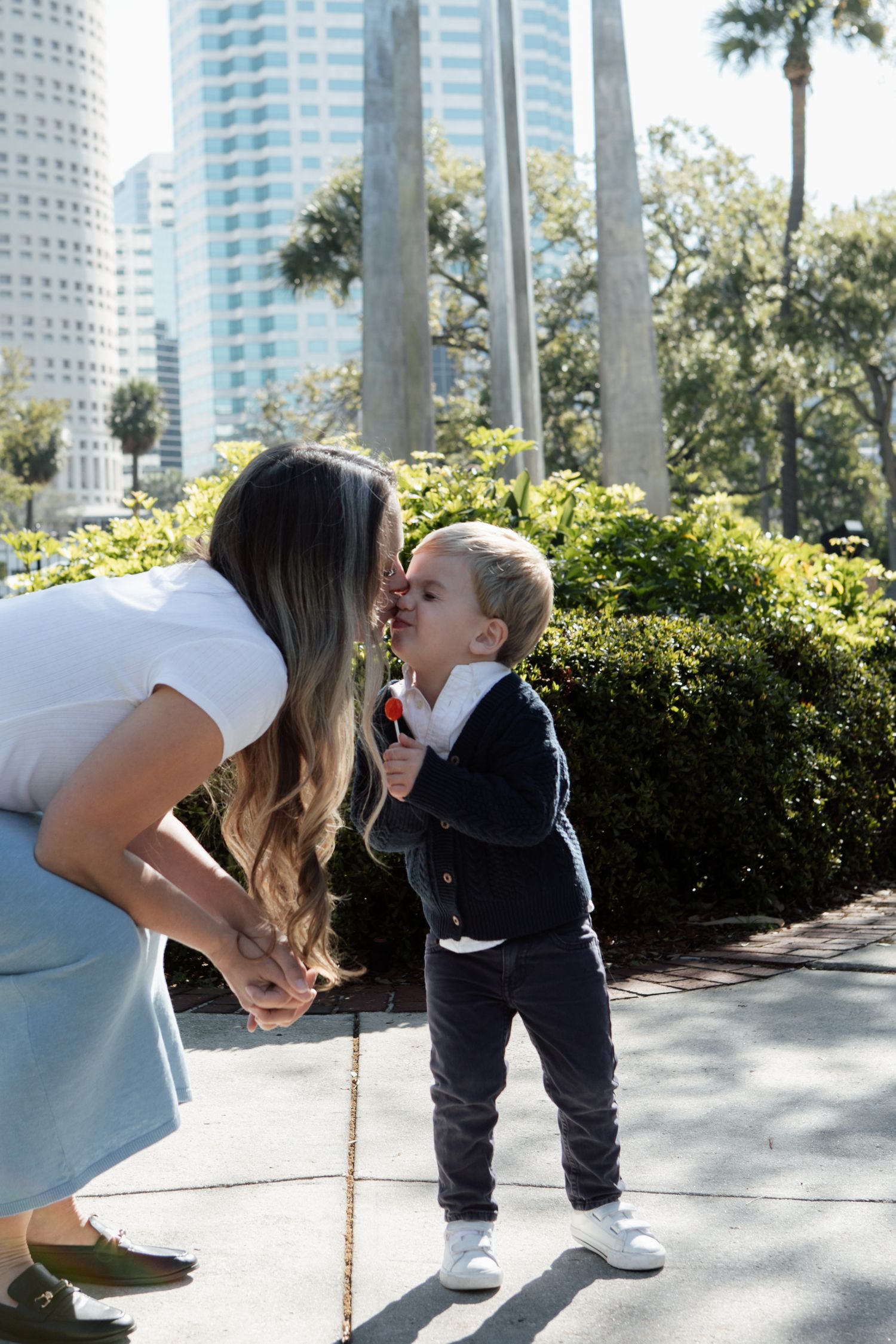 Mom shares a sweet kiss with her young son holding a lollipop in a sunny city park.